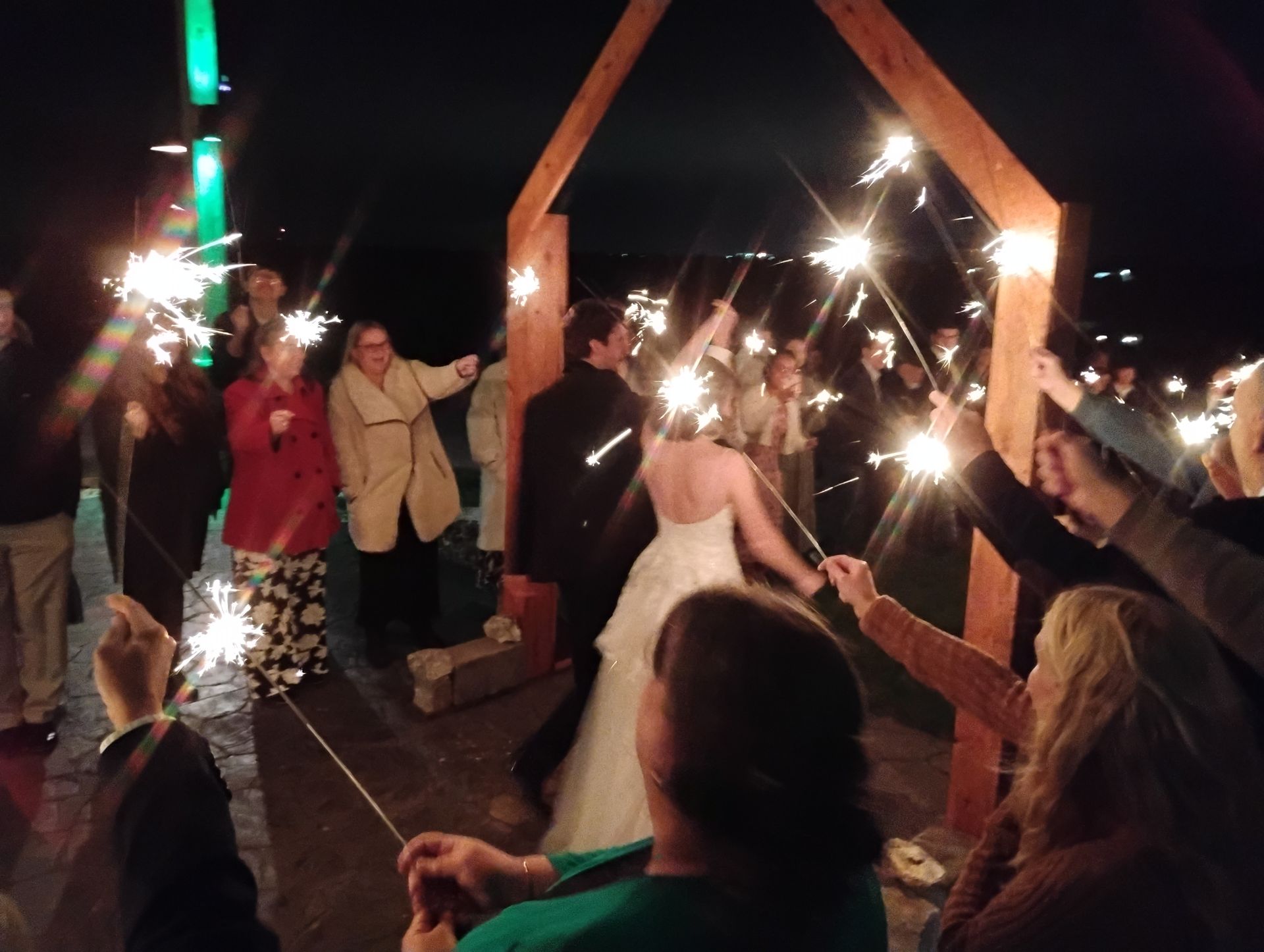 Wedding couple holding sparklers under a wooden arch at night, surrounded by guests with raised sparklers.