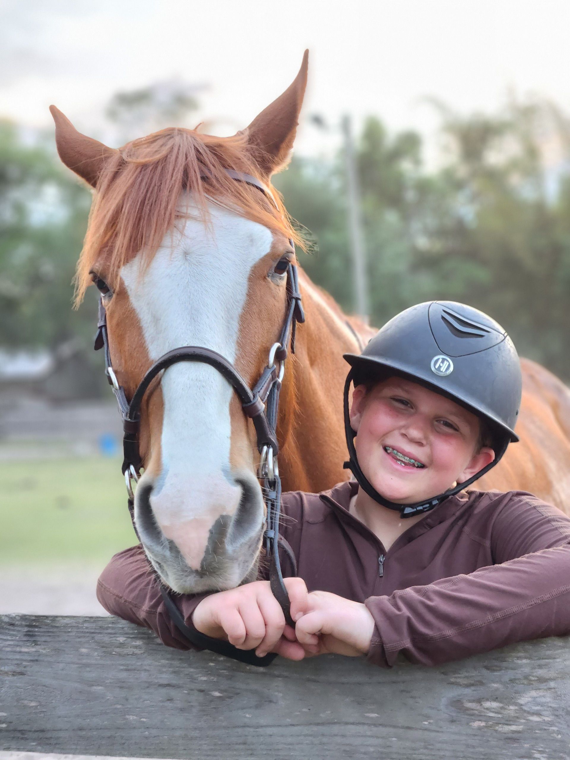 A young boy wearing a helmet stands next to a brown horse
