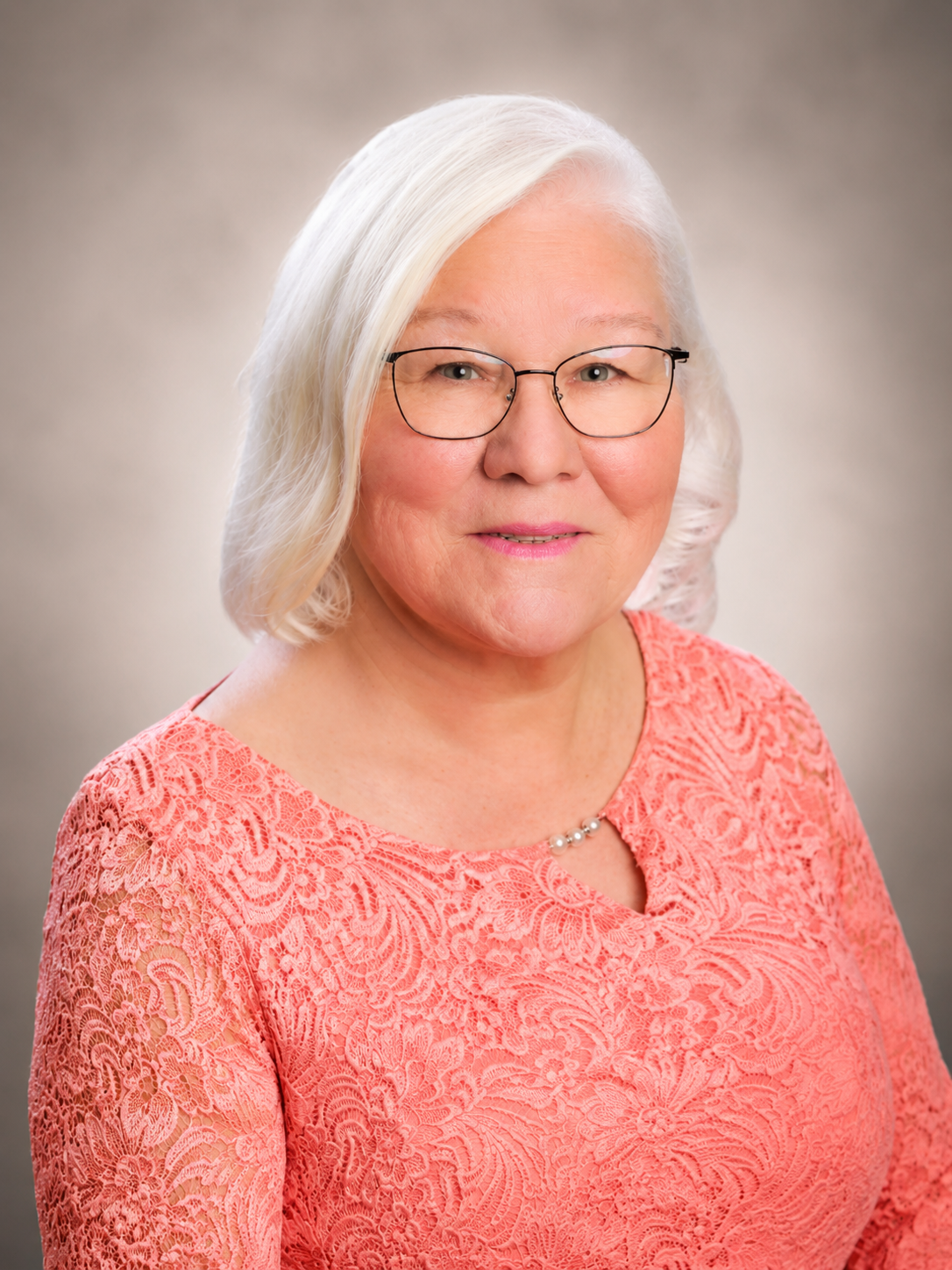 Portrait of a person with white hair and glasses wearing a coral lace top on a neutral background