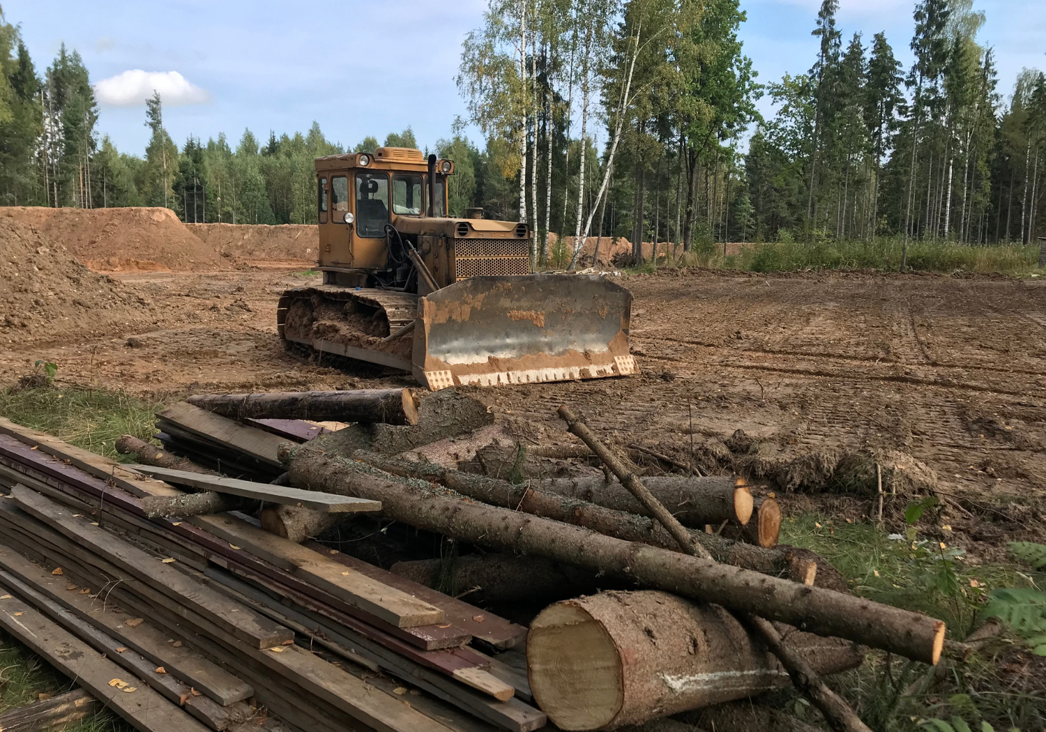 Bulldozer leveling dirt near a pile of logs and boards in a clearing surrounded by trees.