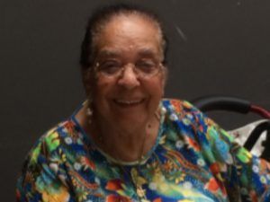 An elderly woman wearing glasses and a colorful shirt is smiling for the camera.