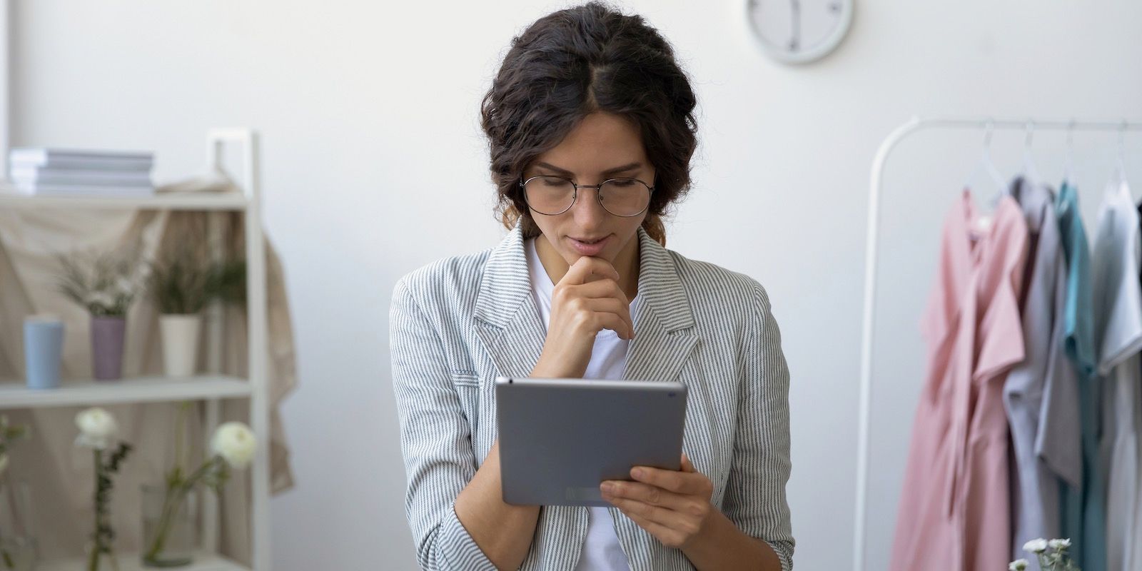 Woman wearing glasses and a striped jacket looks at a tablet, clothes rack in background.