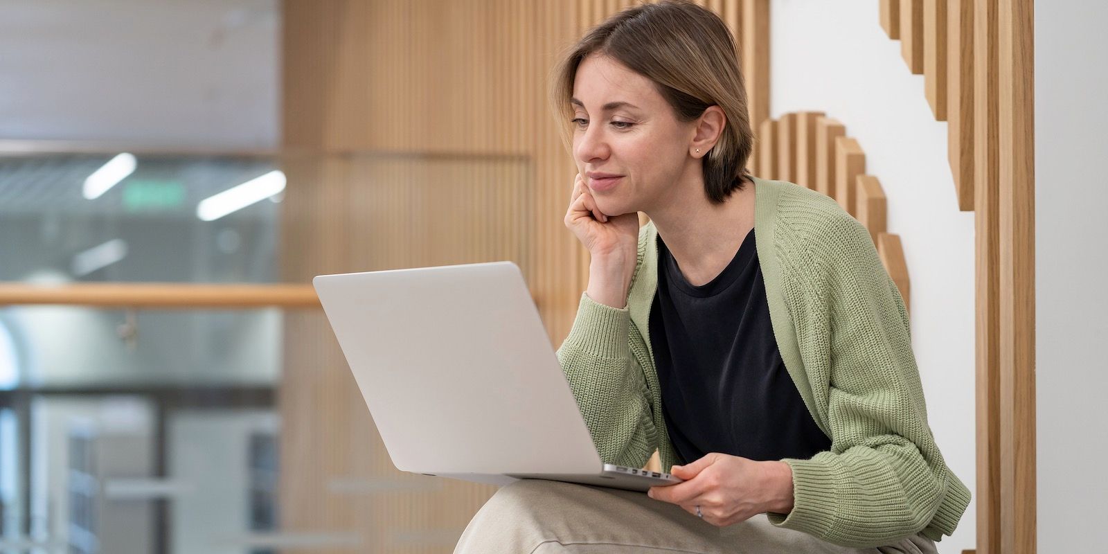 Woman in green cardigan smiles at laptop in a modern, wooden-paneled space.