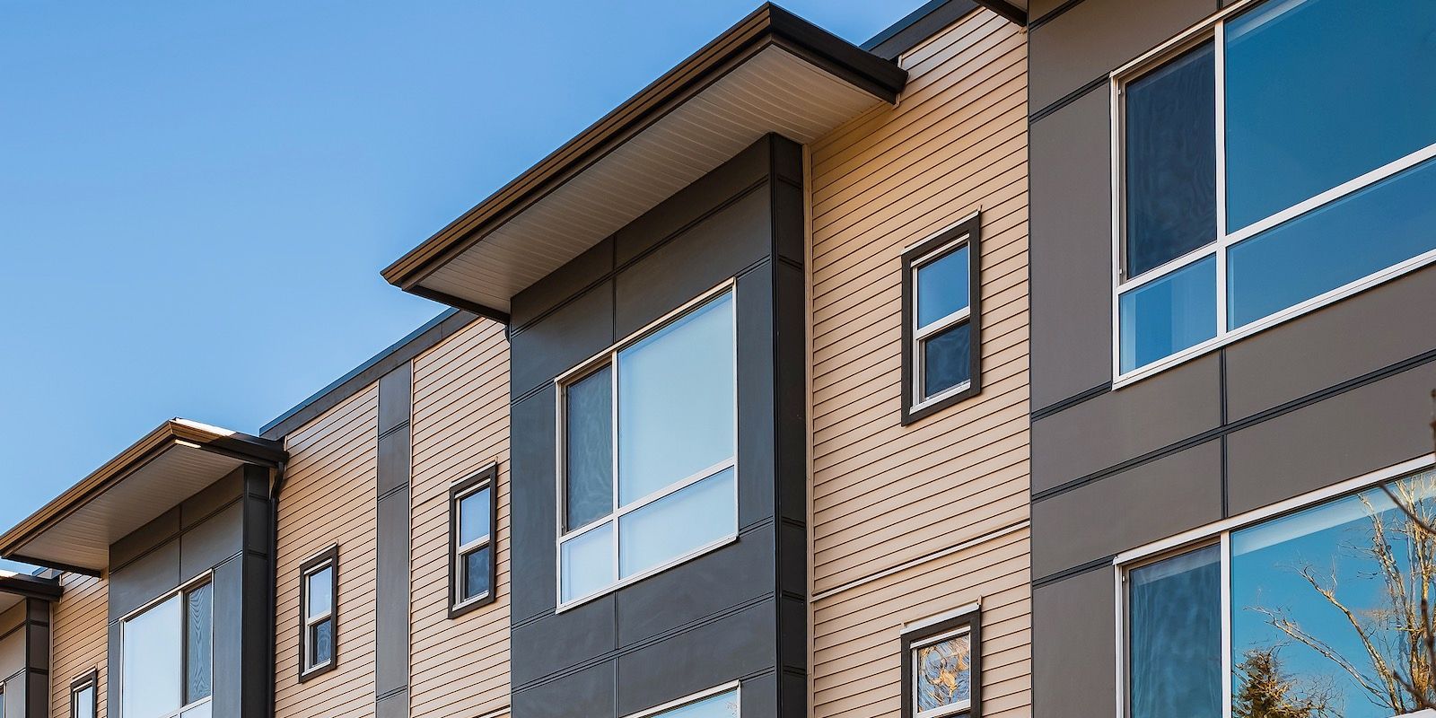 Modern multi-unit housing facade with gray and tan siding, large windows, and a blue sky.