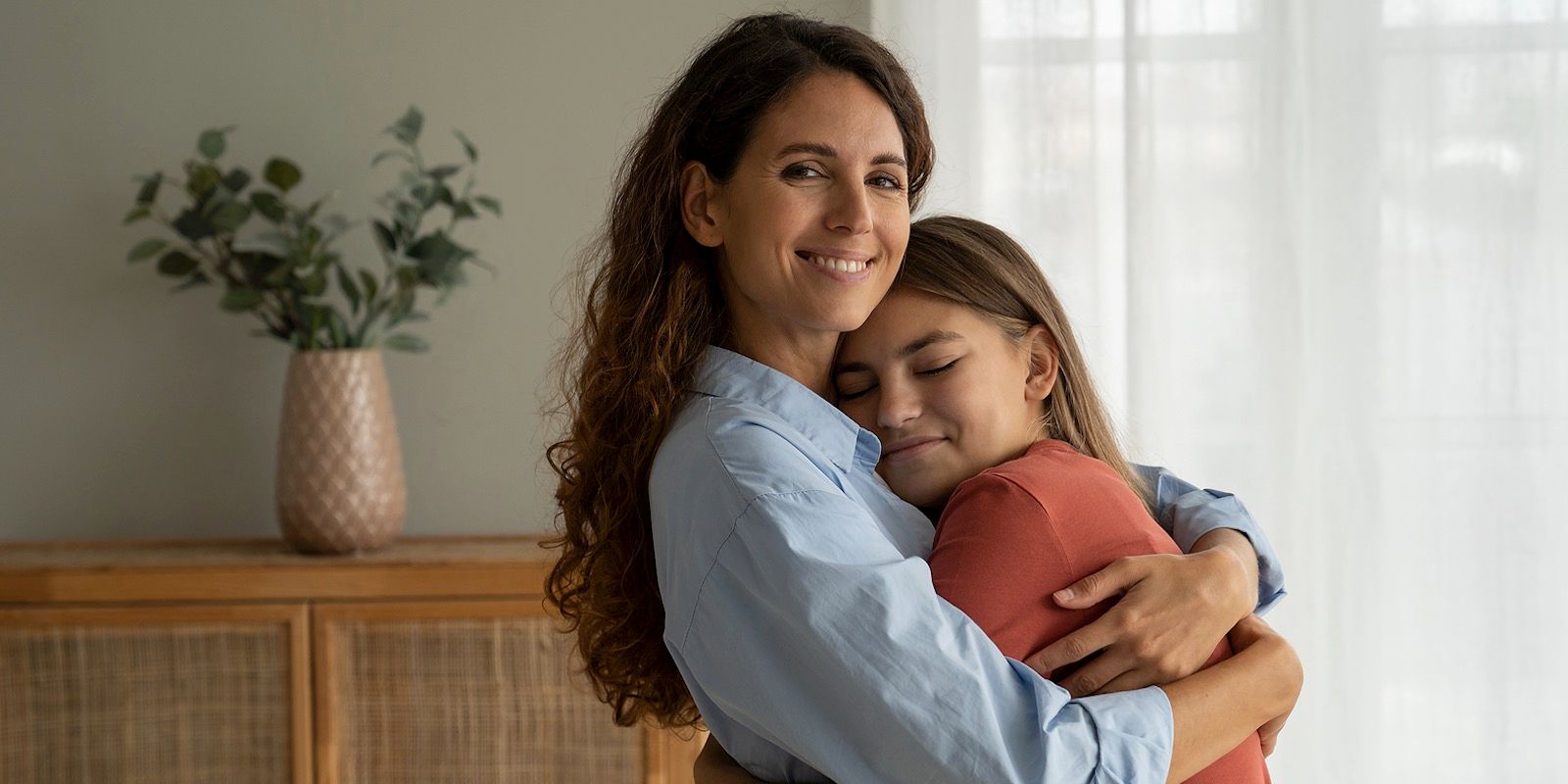 Woman hugging a girl; both are smiling. Indoors with a neutral background.