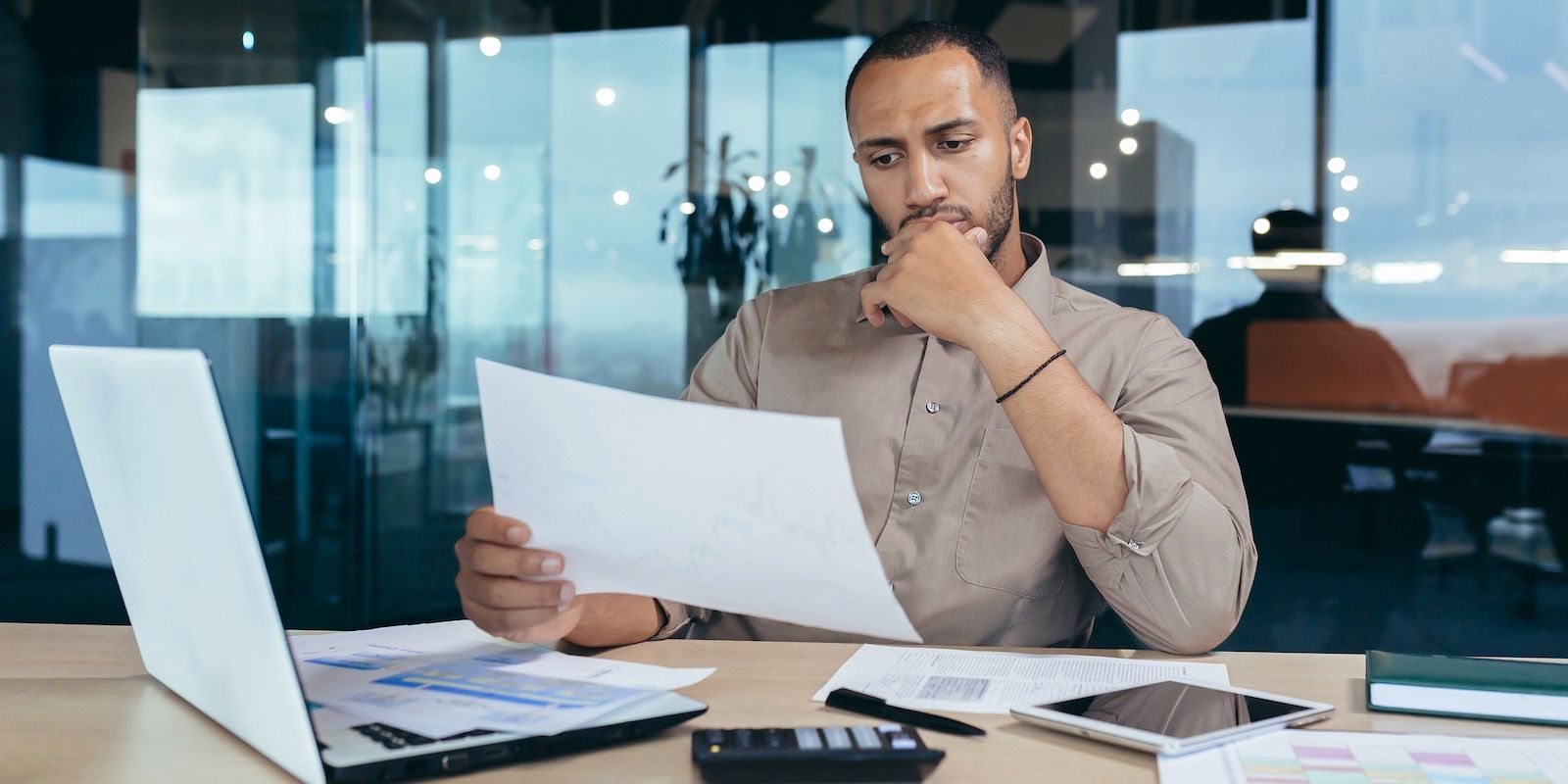 A man in a tan shirt sits at a desk, looking at documents. He appears thoughtful.