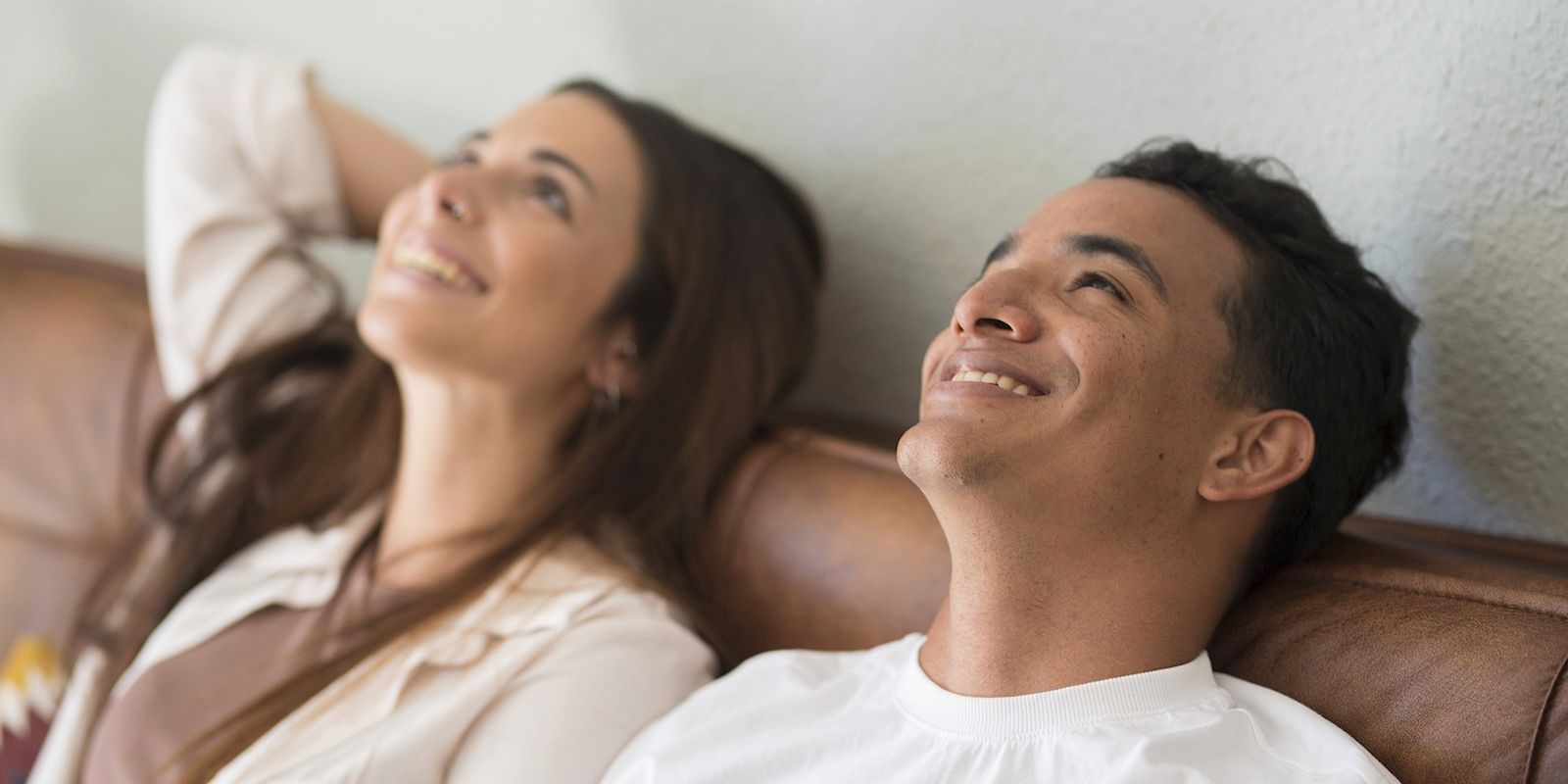 A couple smiling while looking upward, relaxing on a brown couch. The woman has her arms behind her head.