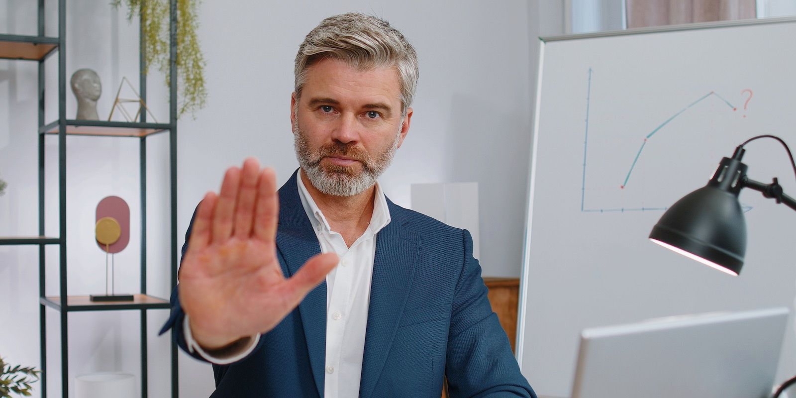 A man in a suit is holding up his hand in a stop gesture. Office setting with whiteboard and lamp.