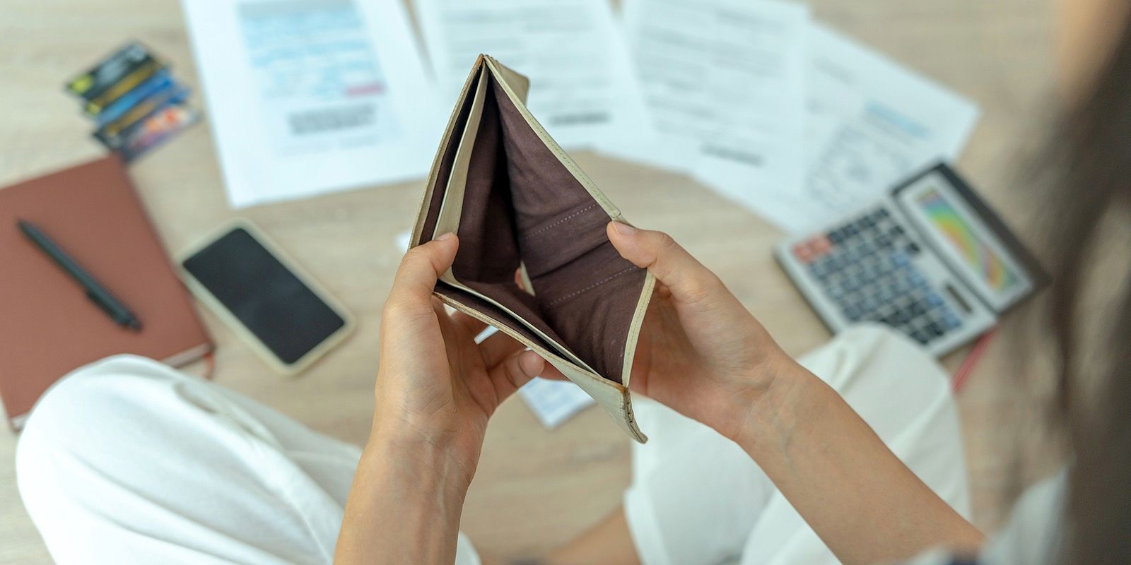 Person holds an empty wallet, surrounded by bills, notebook, phone and calculator, depicting financial hardship.