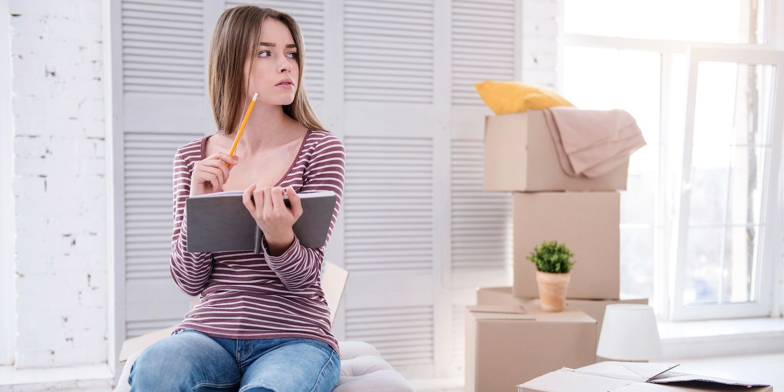 Woman with notebook and pencil, looking thoughtful while surrounded by moving boxes.