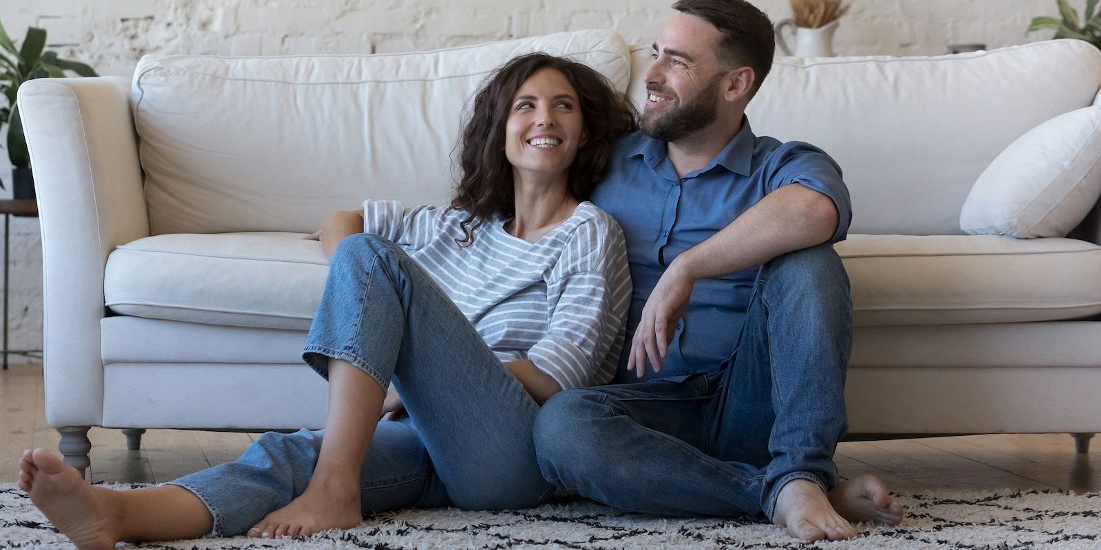 A smiling couple sits on the floor in front of a couch. They are relaxed and look happy.