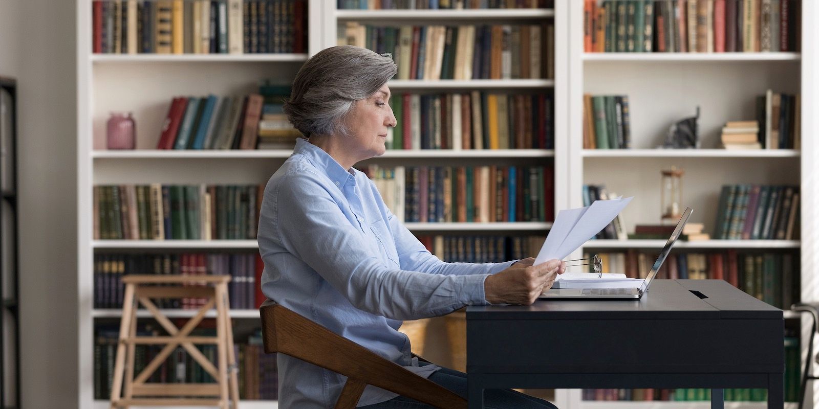 An older woman in a blue shirt sits at a desk with a laptop and papers in front of a bookcase.
