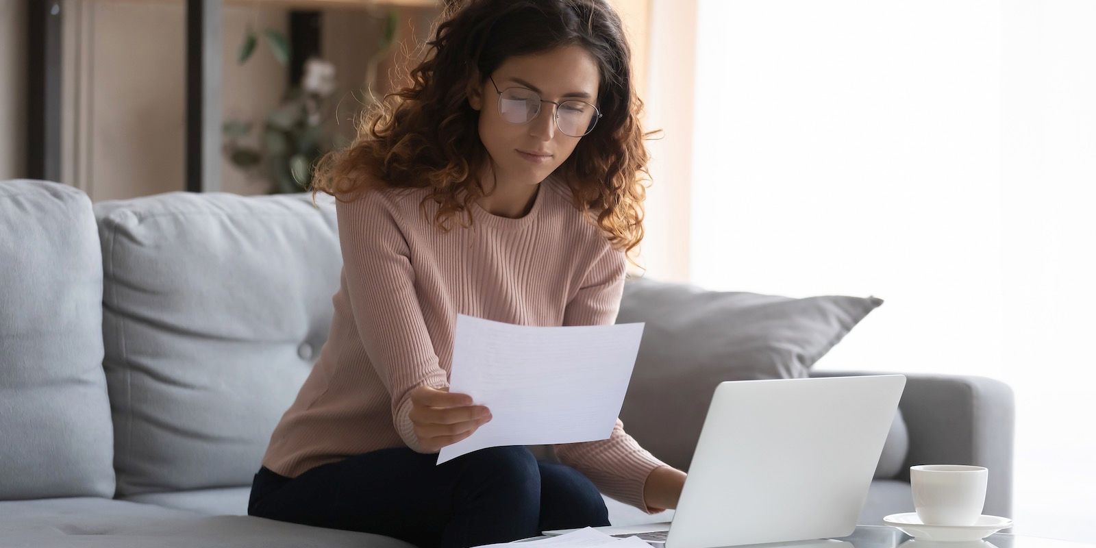 Woman in glasses reviews documents while working on a laptop, sitting on a sofa.