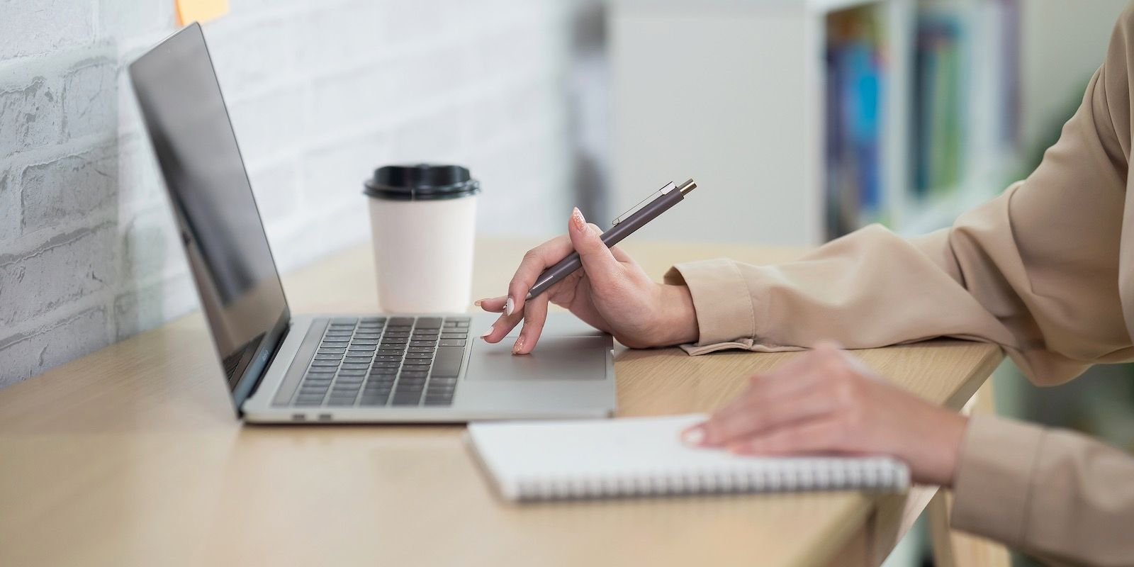 Person working on laptop with a pen and notebook at a desk, near a coffee cup.