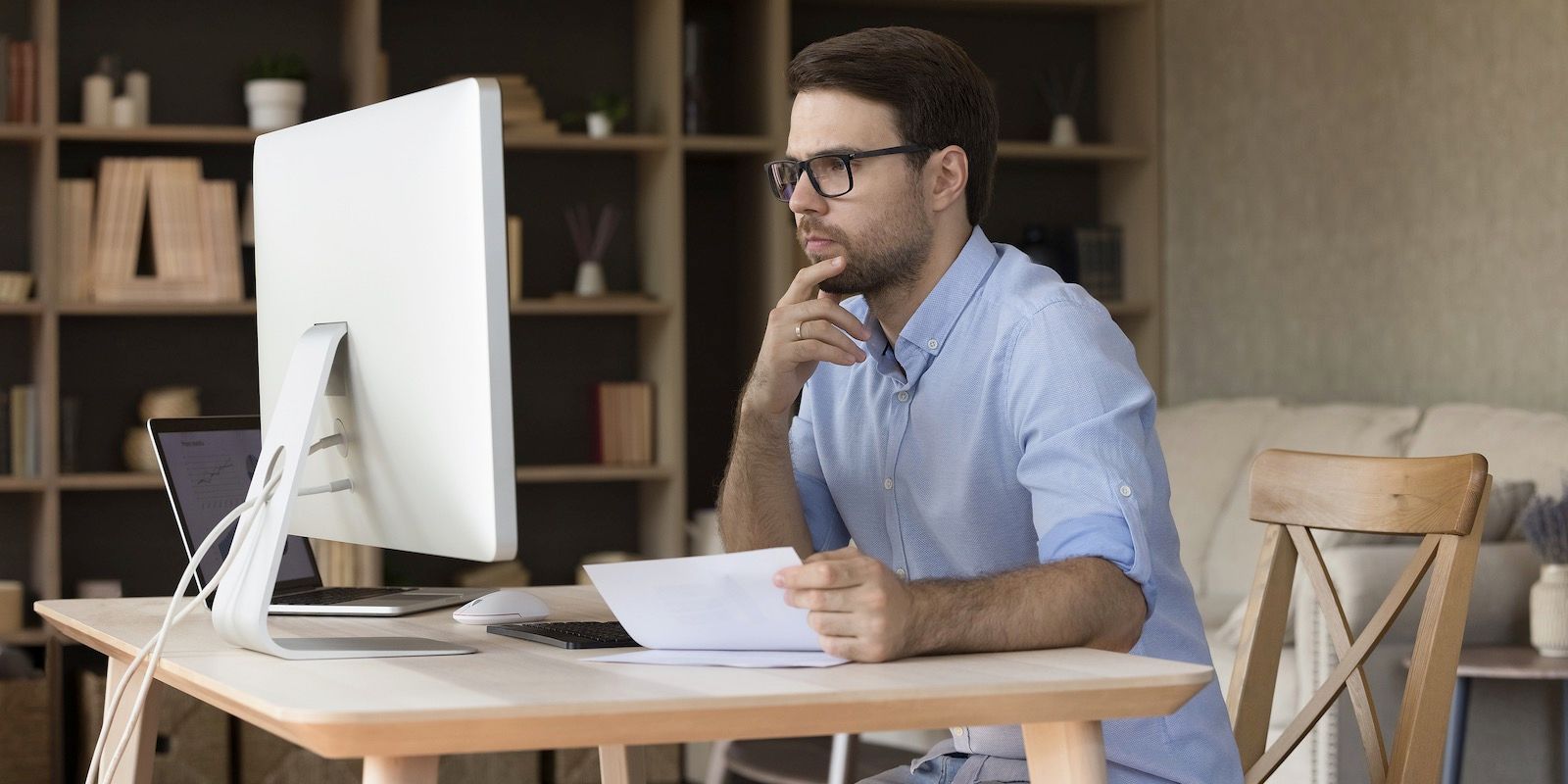 Man wearing glasses at a desk with a computer, looking thoughtful and holding papers.