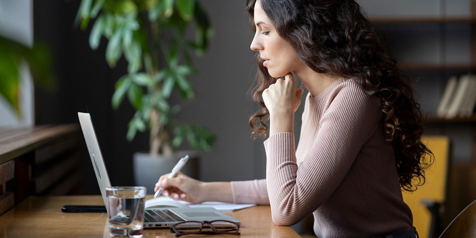Woman with curly hair, writing at a laptop, resting her chin. A glass of water sits on the desk.