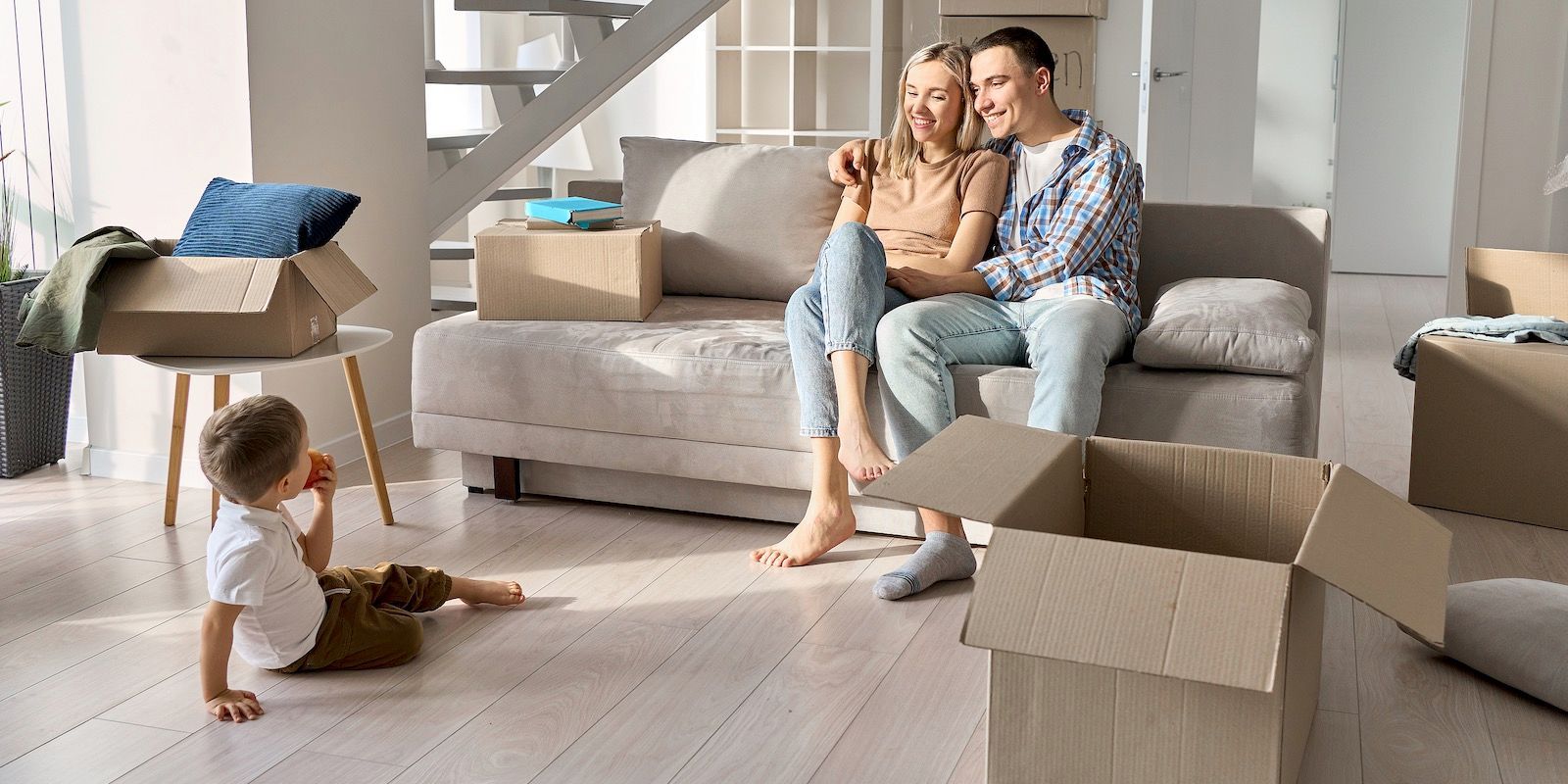 A family in a new home with boxes. The couple sits on a couch, hugging, while their child plays on the floor.
