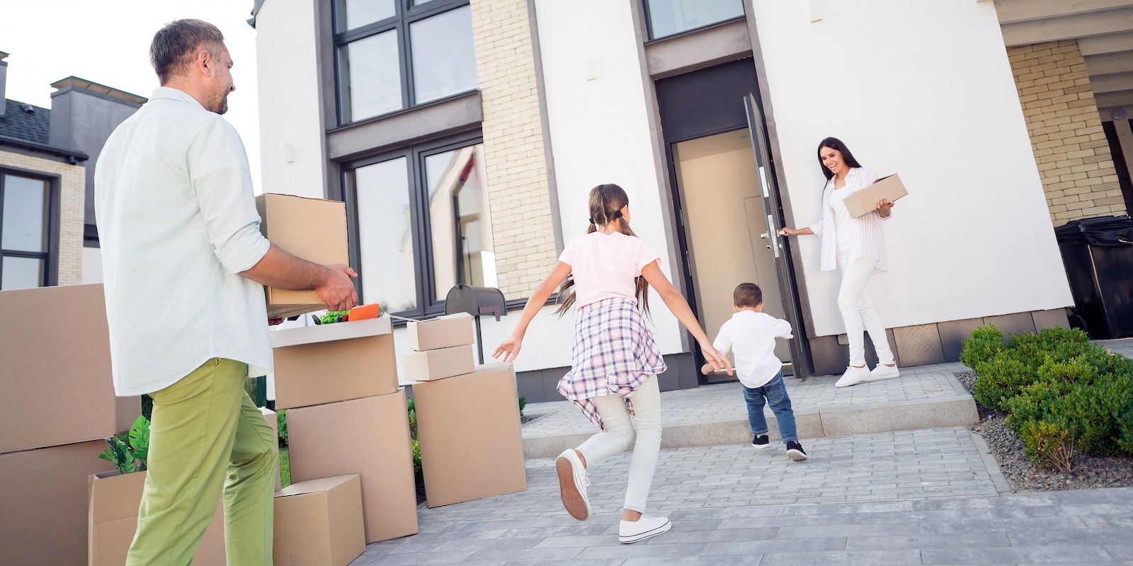 Family moving into a new house: parents and children. Boxes, running kids, open door, stone walkway, white house.