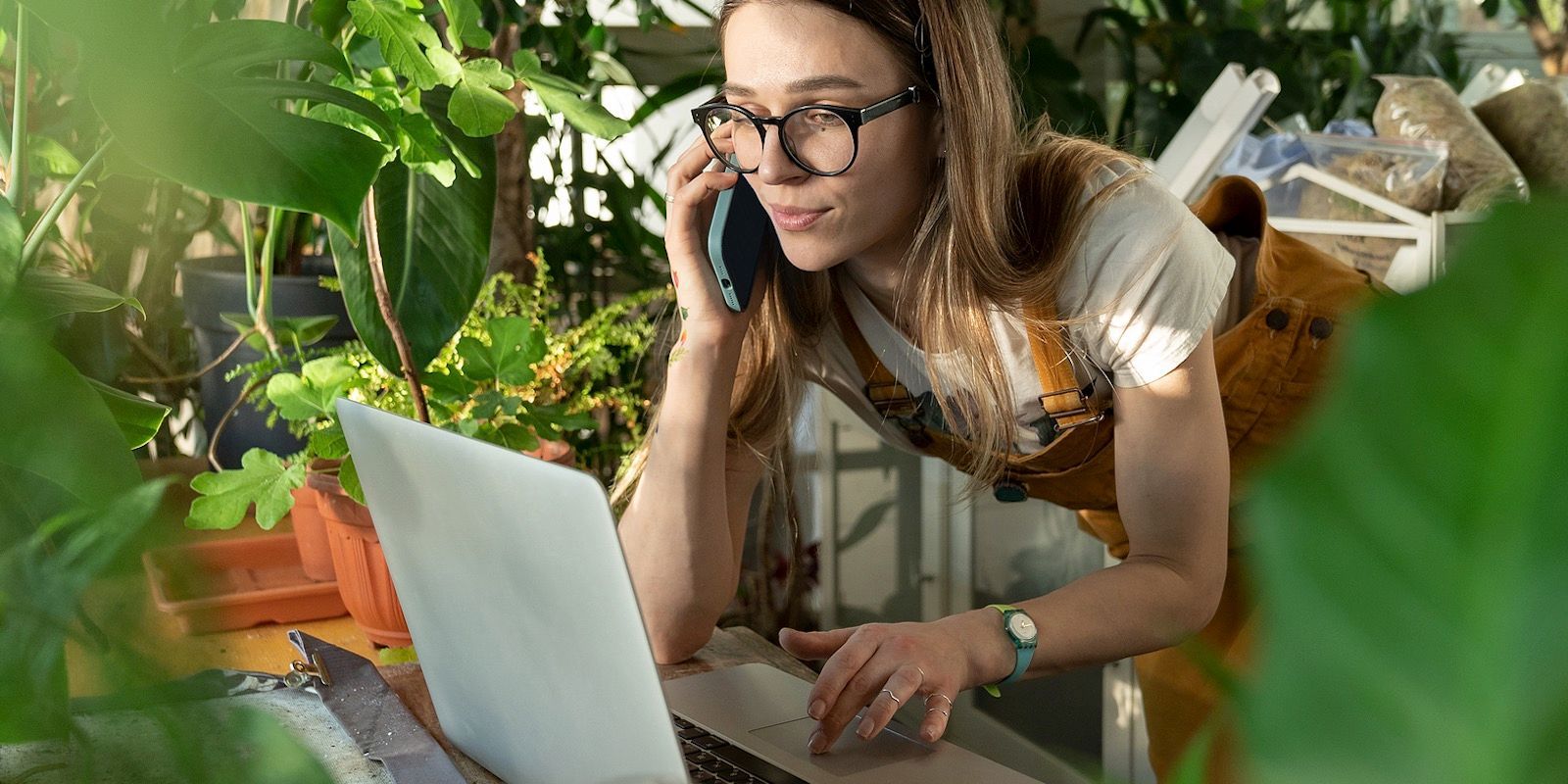 Woman with glasses on a phone call using a laptop in a greenhouse, surrounded by plants.