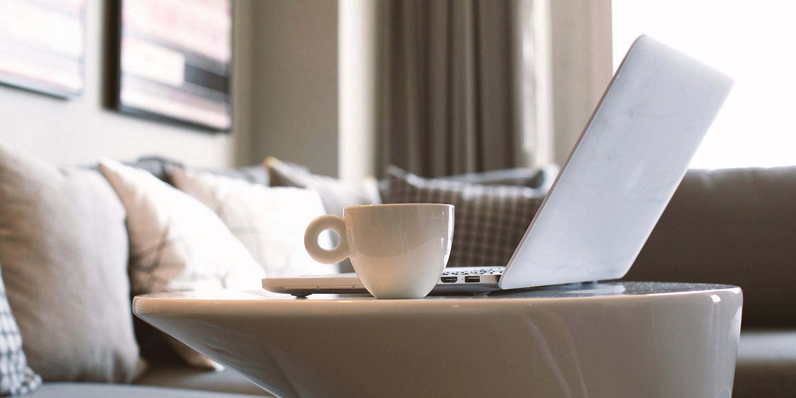 Laptop and coffee cup on a white table in front of a sofa. Interior shot, soft lighting.
