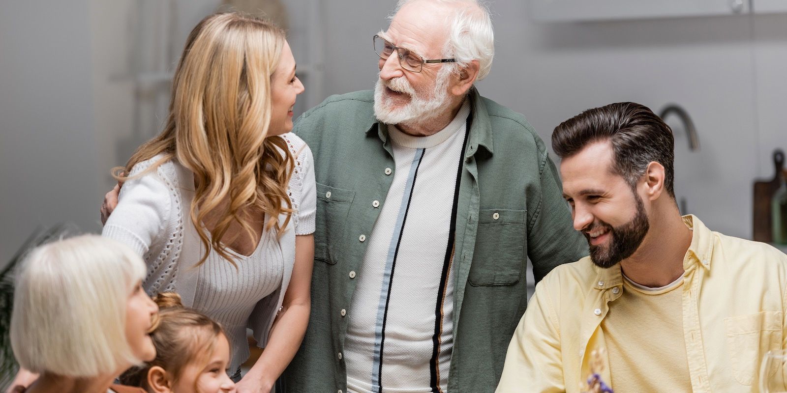 A family of five smiling in a kitchen. A senior man with glasses looks at a woman with long blonde hair.