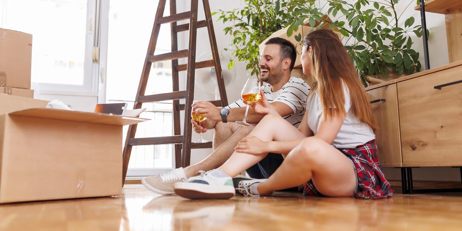 Couple toasting drinks while sitting on floor in new home, near boxes and ladder.