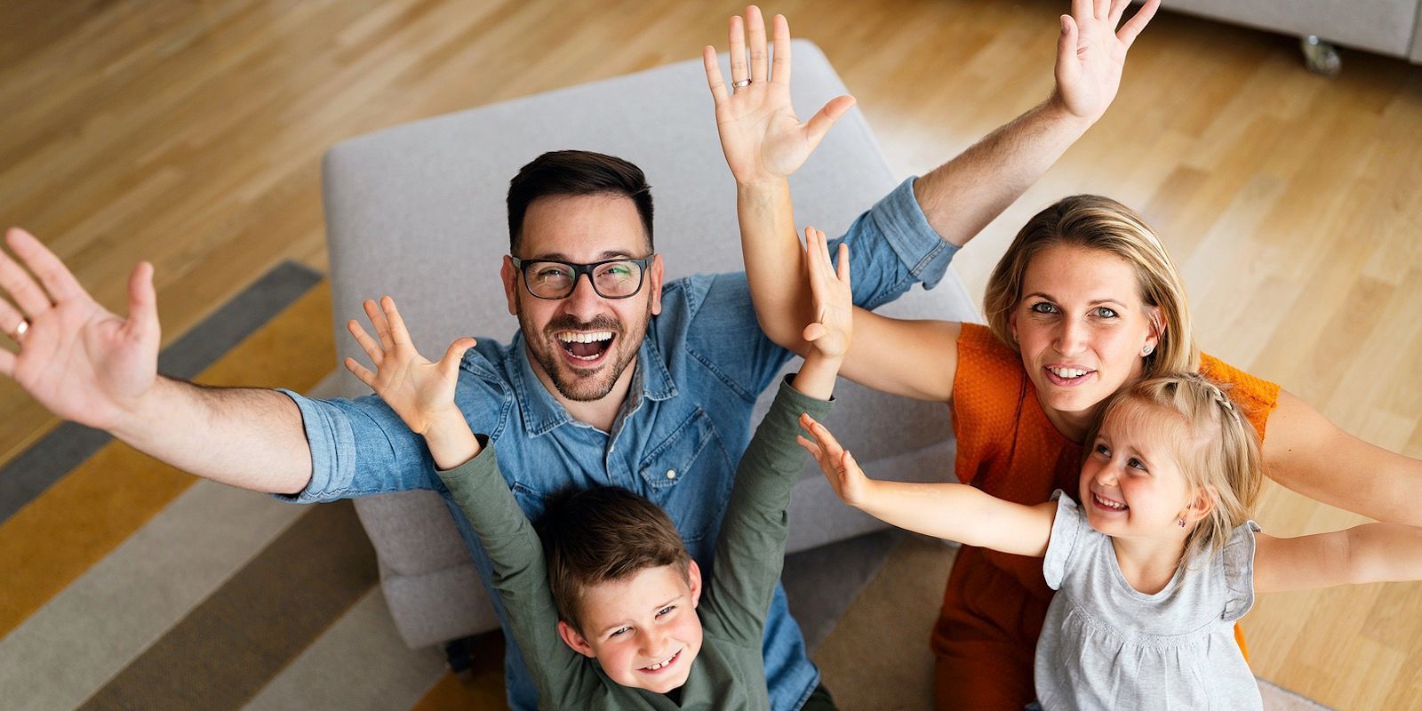 Family of four, smiling with arms raised, sitting on a patterned rug and wooden floor.