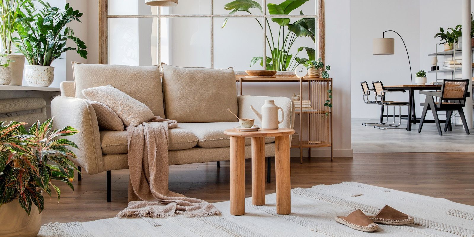 Cozy living room with beige sofa, wooden table, rug, and plants. Dining area visible in the background.