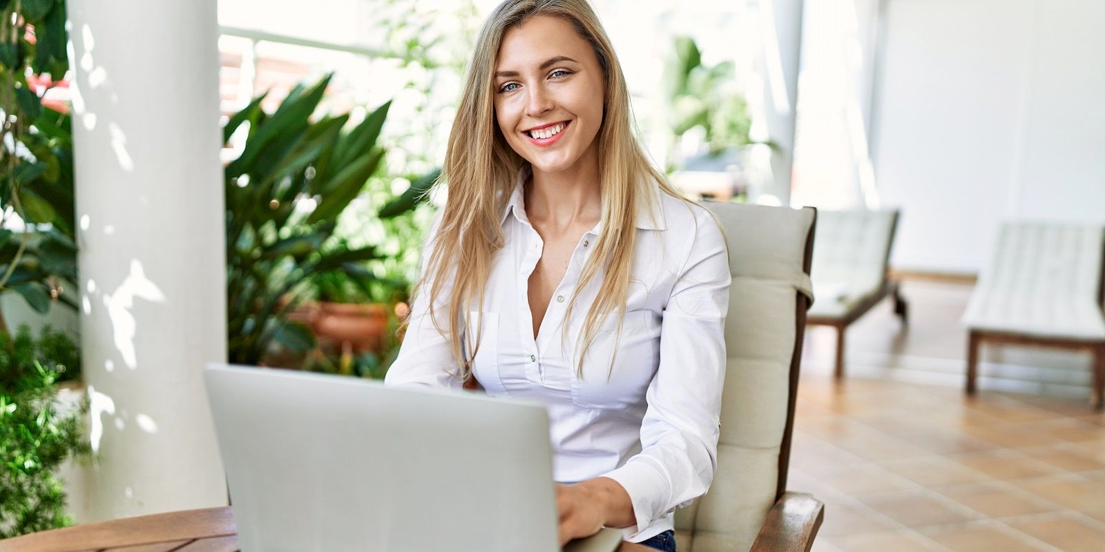 Woman sitting outdoors, smiling, using a laptop, wearing a white shirt.