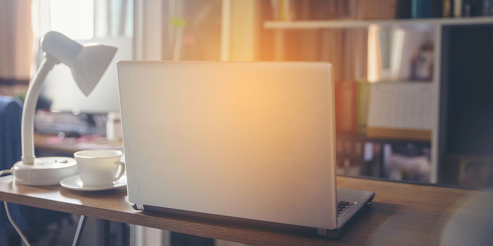Laptop on a wooden desk with a lamp and a cup in a room with a window.
