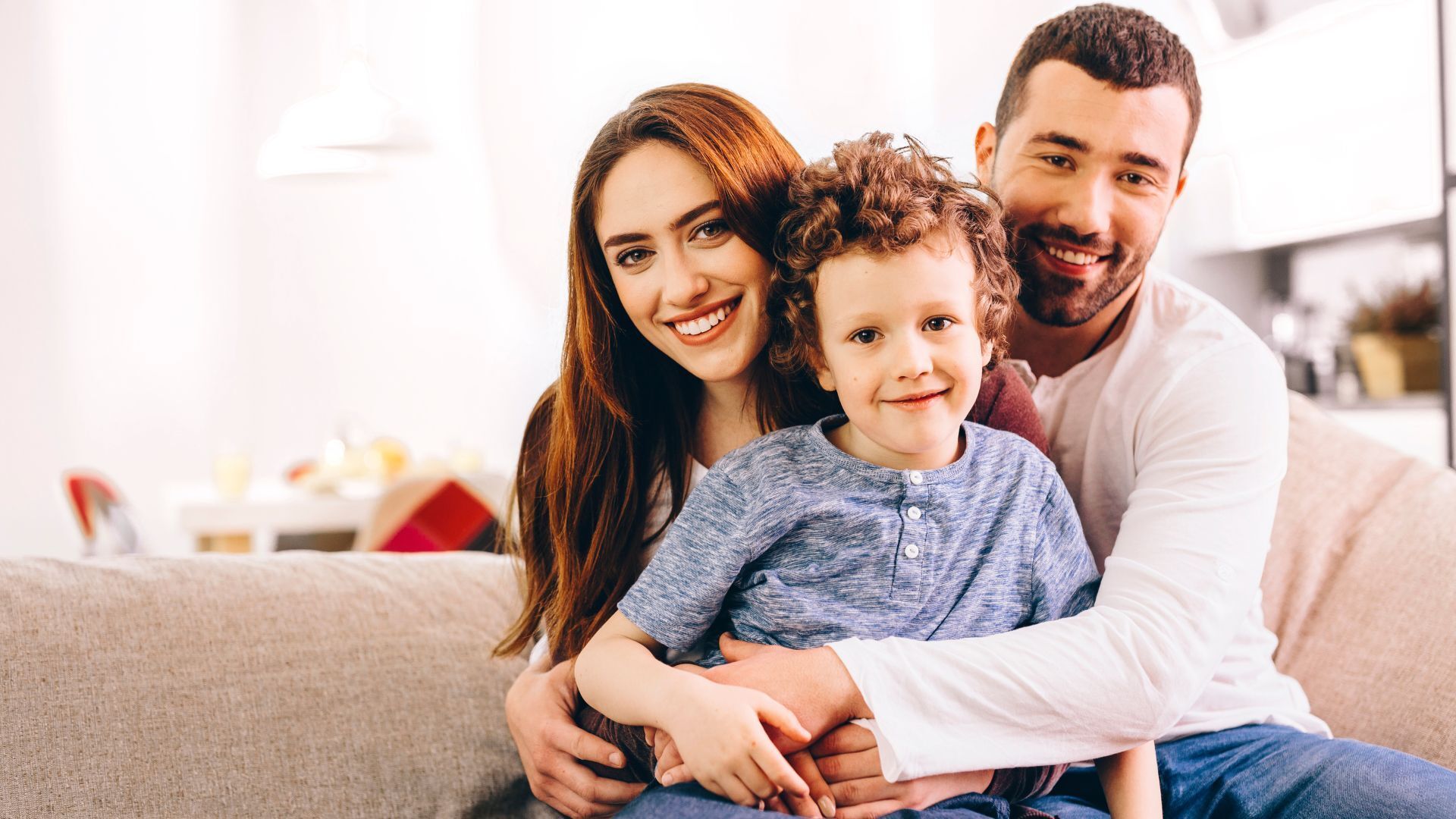 A smiling family of three on a couch: mother, father, and young child.