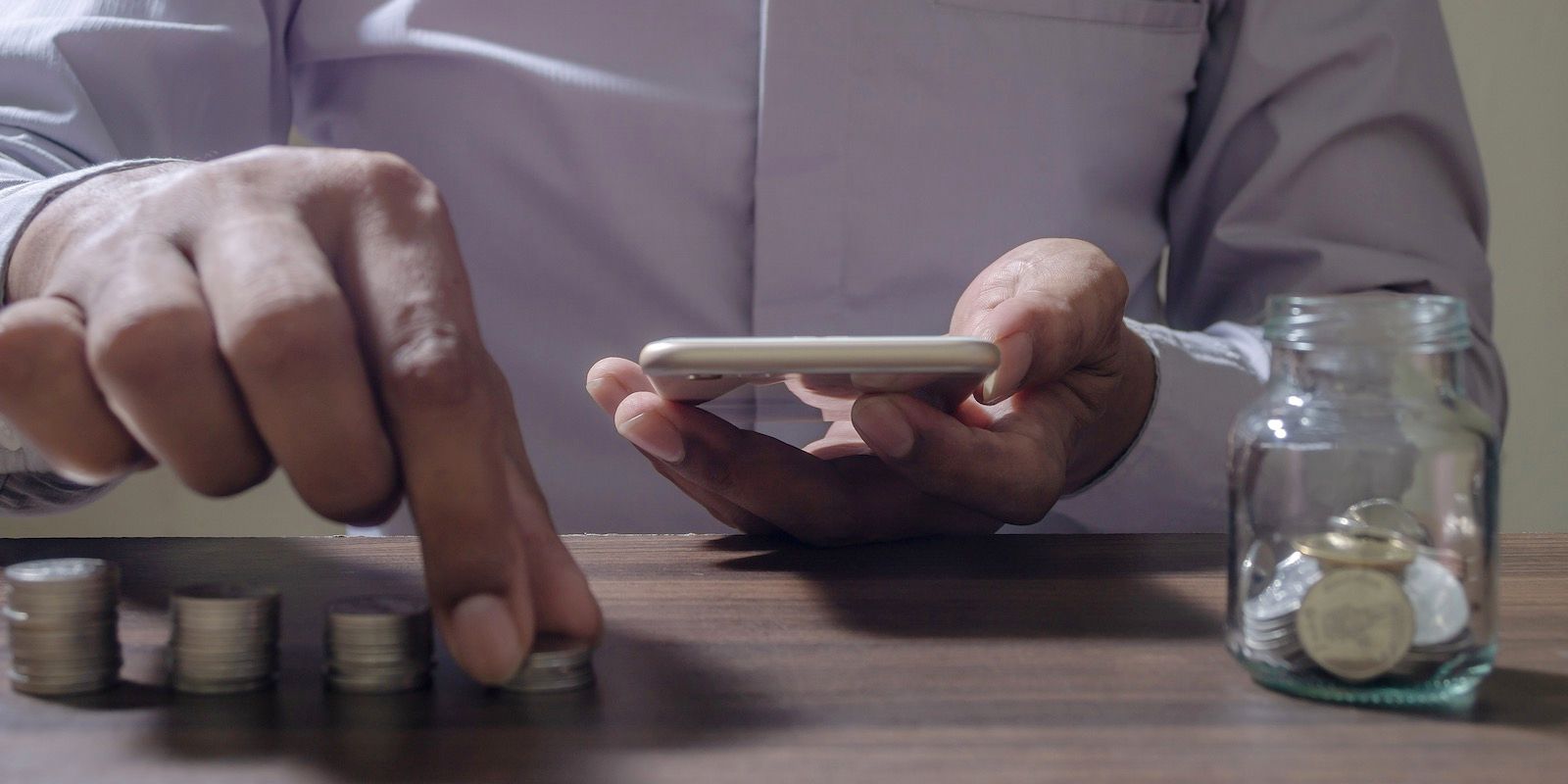 Person counting coins with a smartphone, jar of money on a wood table.