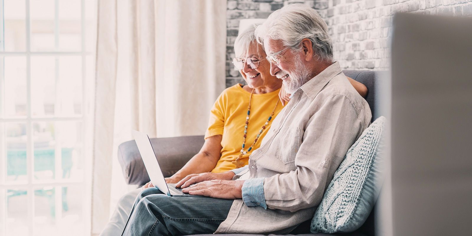 Elderly couple using a laptop on a sofa, near a window. The man is typing while the woman watches.