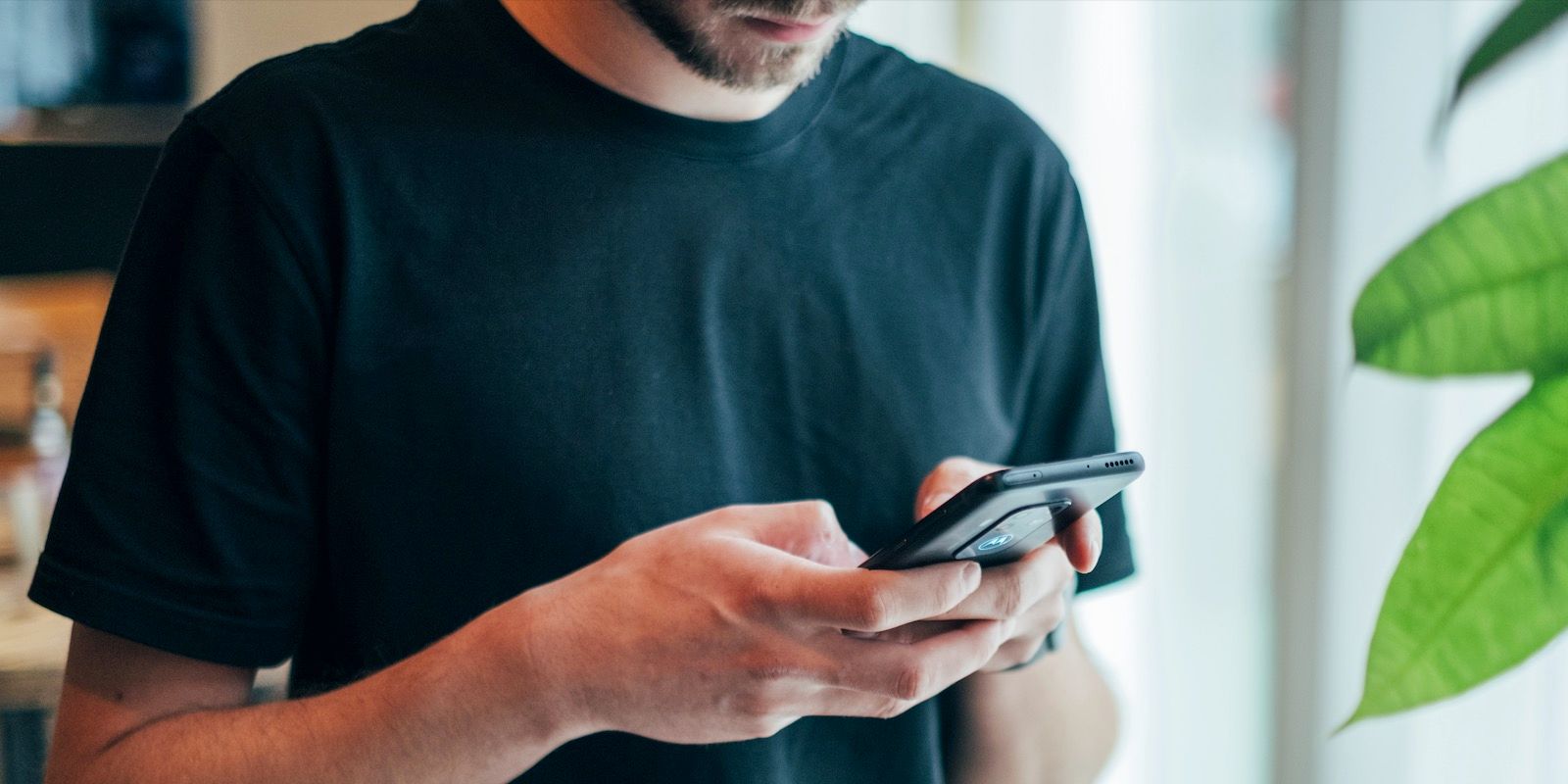 Man in black shirt using a phone, light background, green plant in corner.