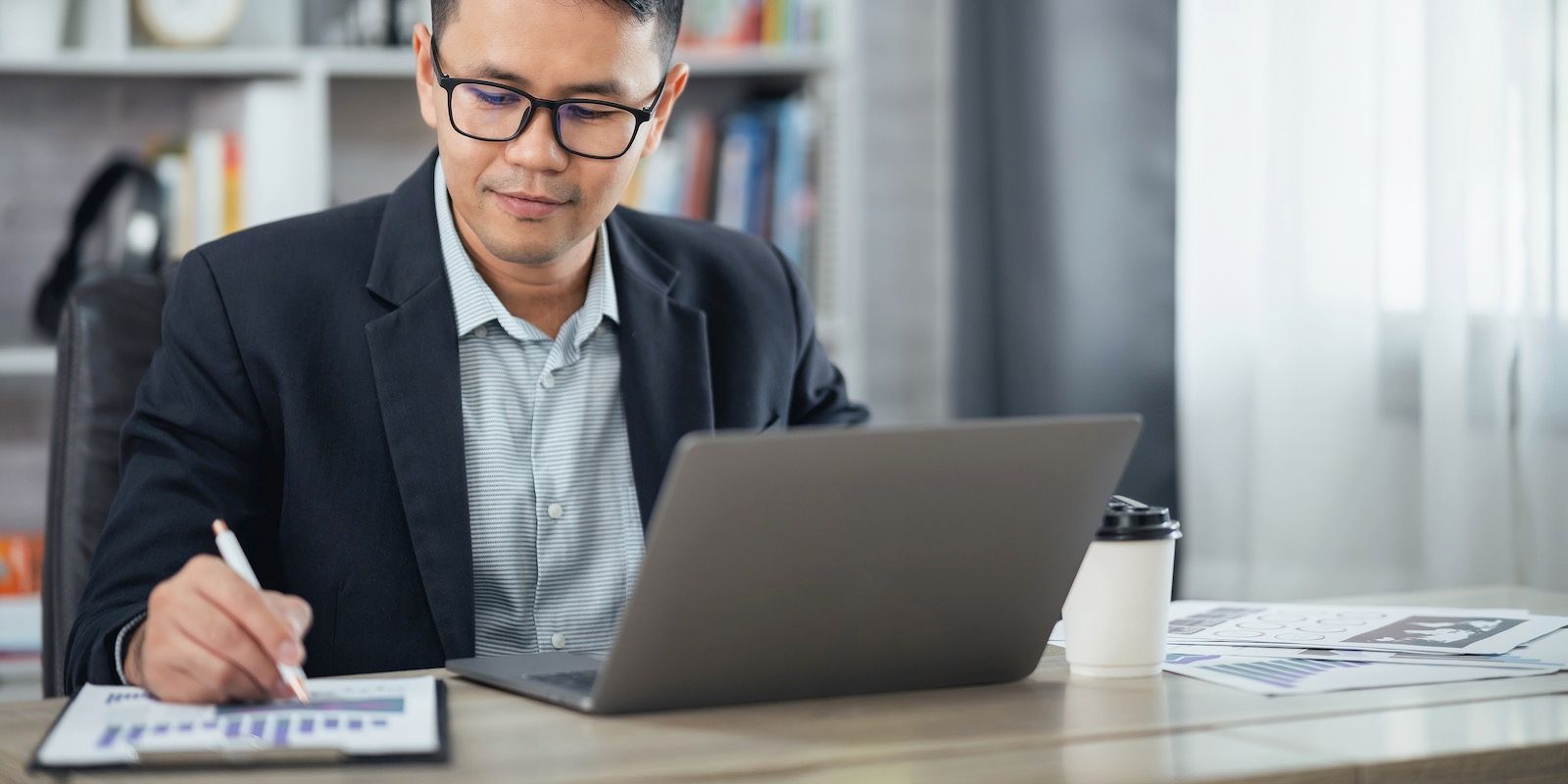A man in glasses and a suit works on a laptop and analyzes a chart in an office setting.