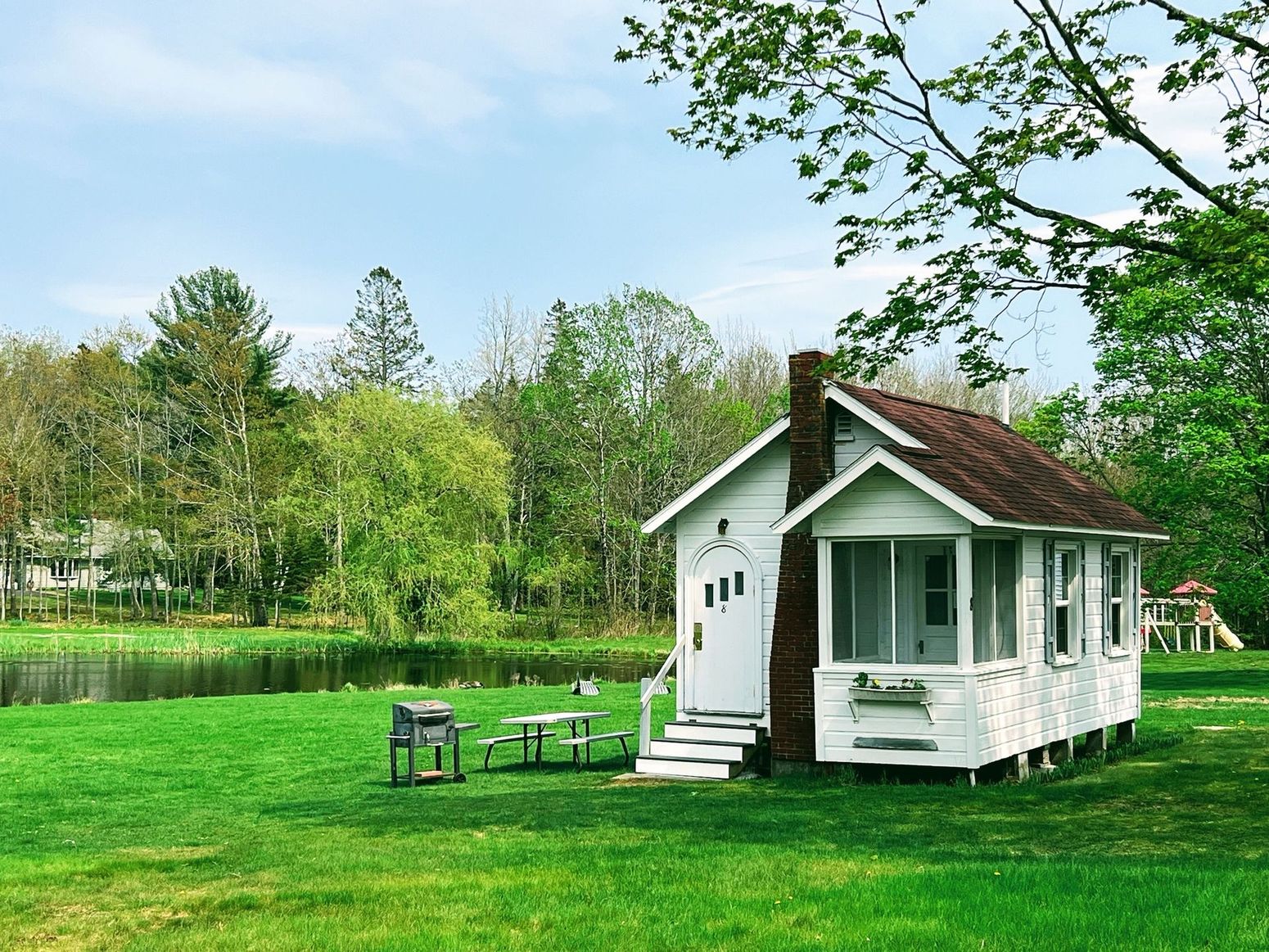 White cottage with a porch sits on a green lawn near trees and a small pond, on a sunny day.