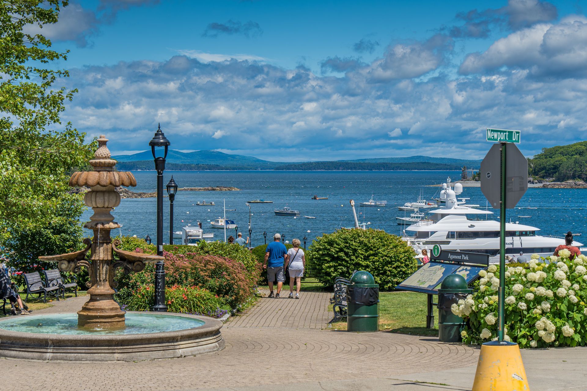 Scenic view of a harbor with fountain, couple walking, boats, and blue water.