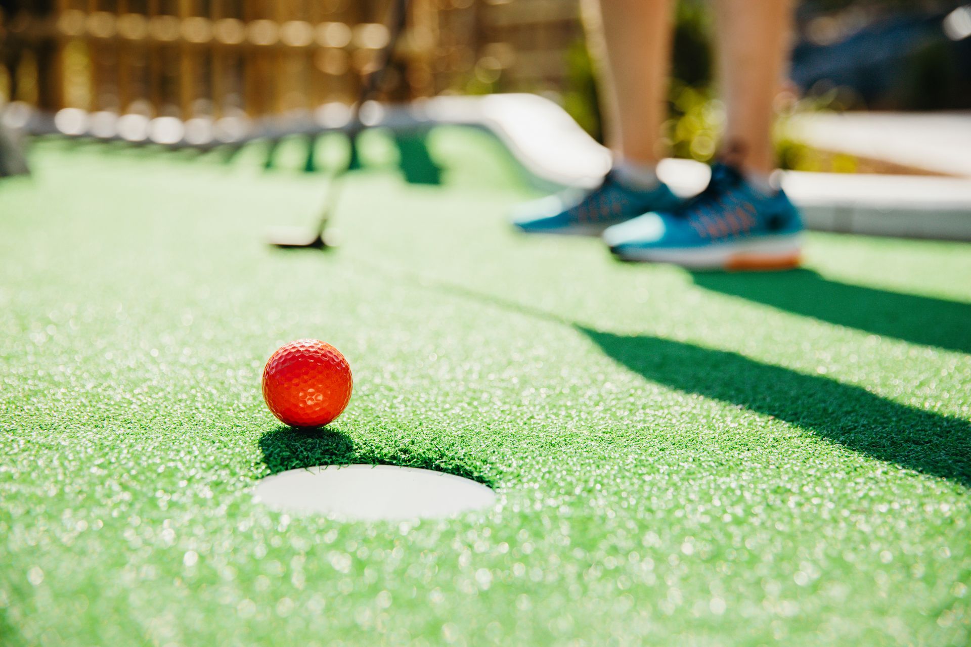 Red mini golf ball near the hole, with a person putting in the background.
