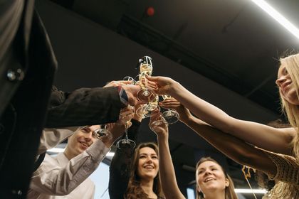 A group of people cheers with champagne flutes held together in a close-up against a dark, festive background.