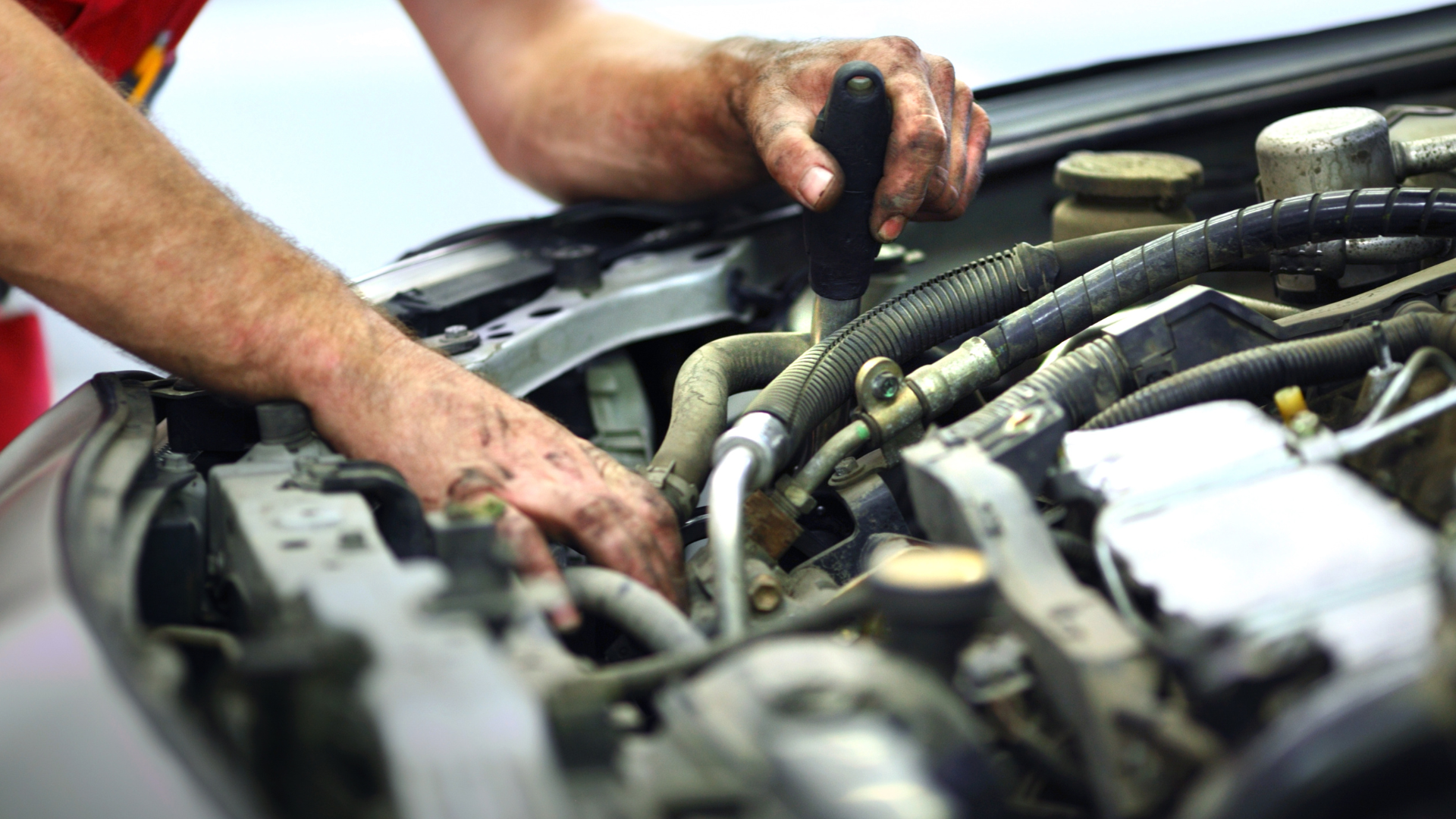 Mechanic working on a car engine, with dirty hands holding a tool, inside the open hood of a vehicle.