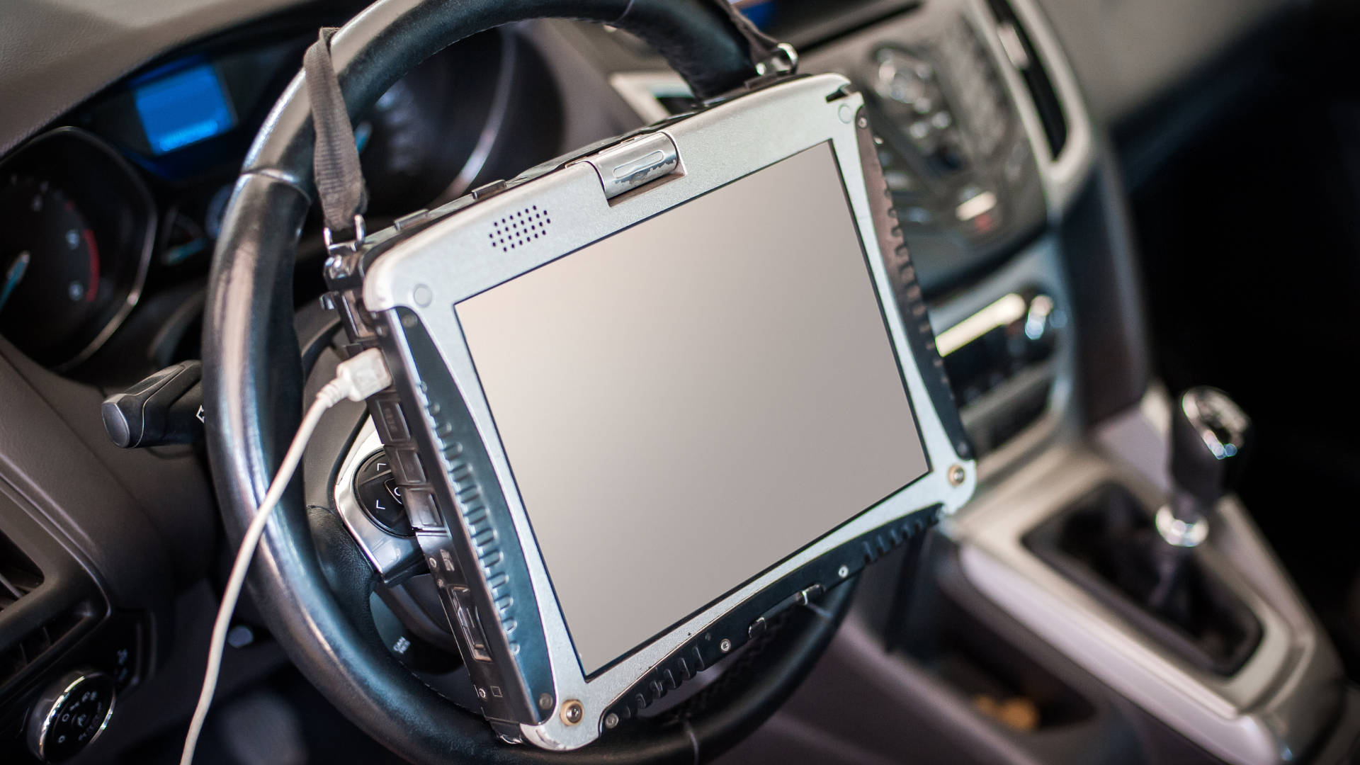 A rugged tablet, likely for diagnostics, is mounted on a car's steering wheel. A cord is plugged into the device.