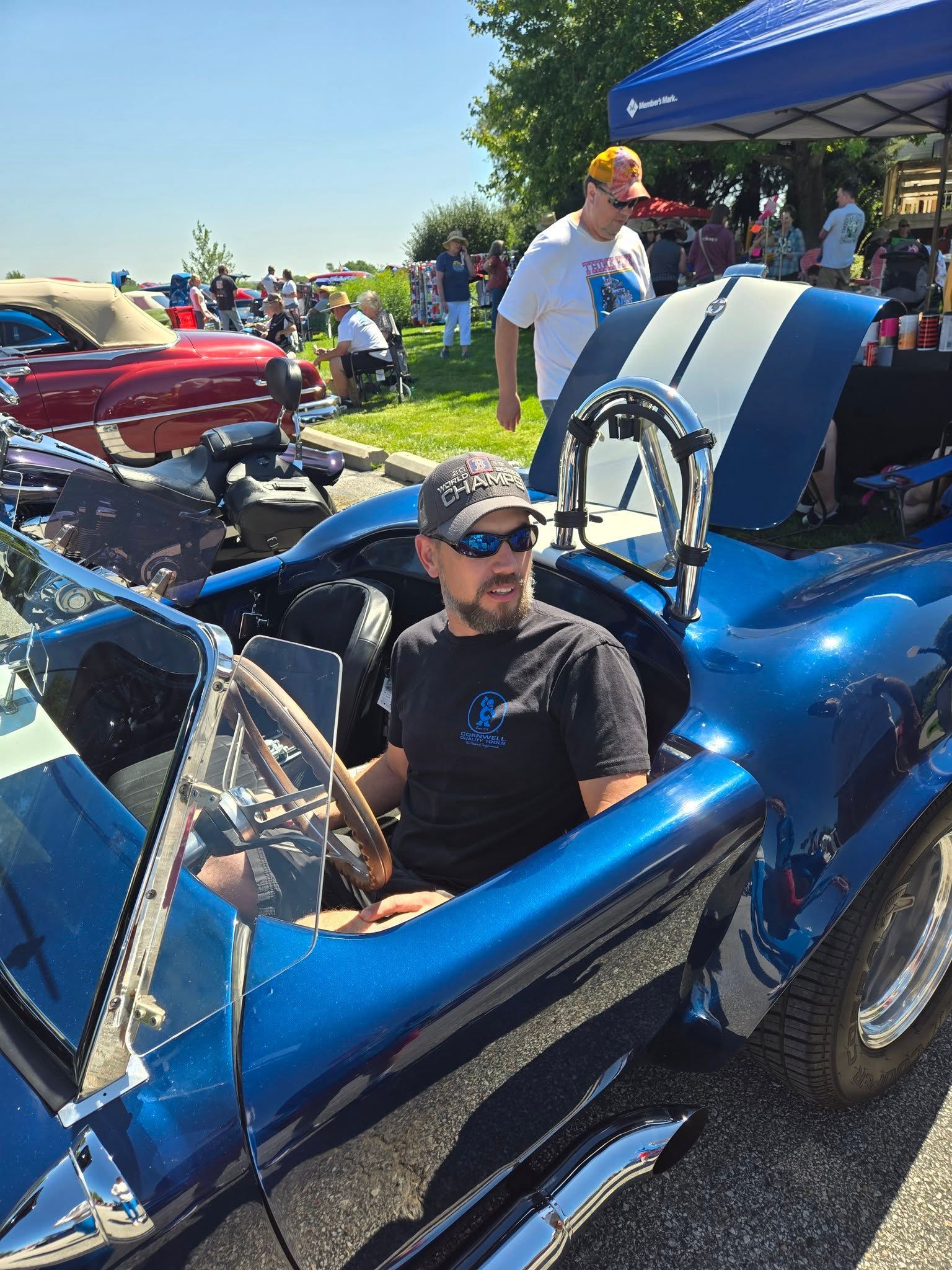 Man sits in a blue classic car with white stripes, at an outdoor car show. Another man walks behind him.