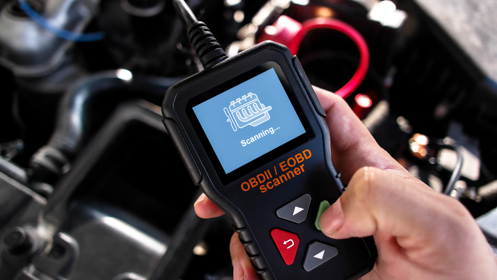 Hand using an OBD2 scanner to diagnose a car engine. The scanner has a screen and buttons, displaying a loading icon.