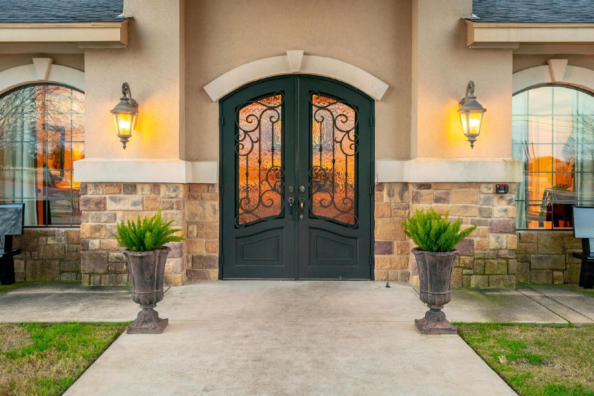 Double black ornate doors, flanked by windows and sconces, lead to a home; two planters sit in front.