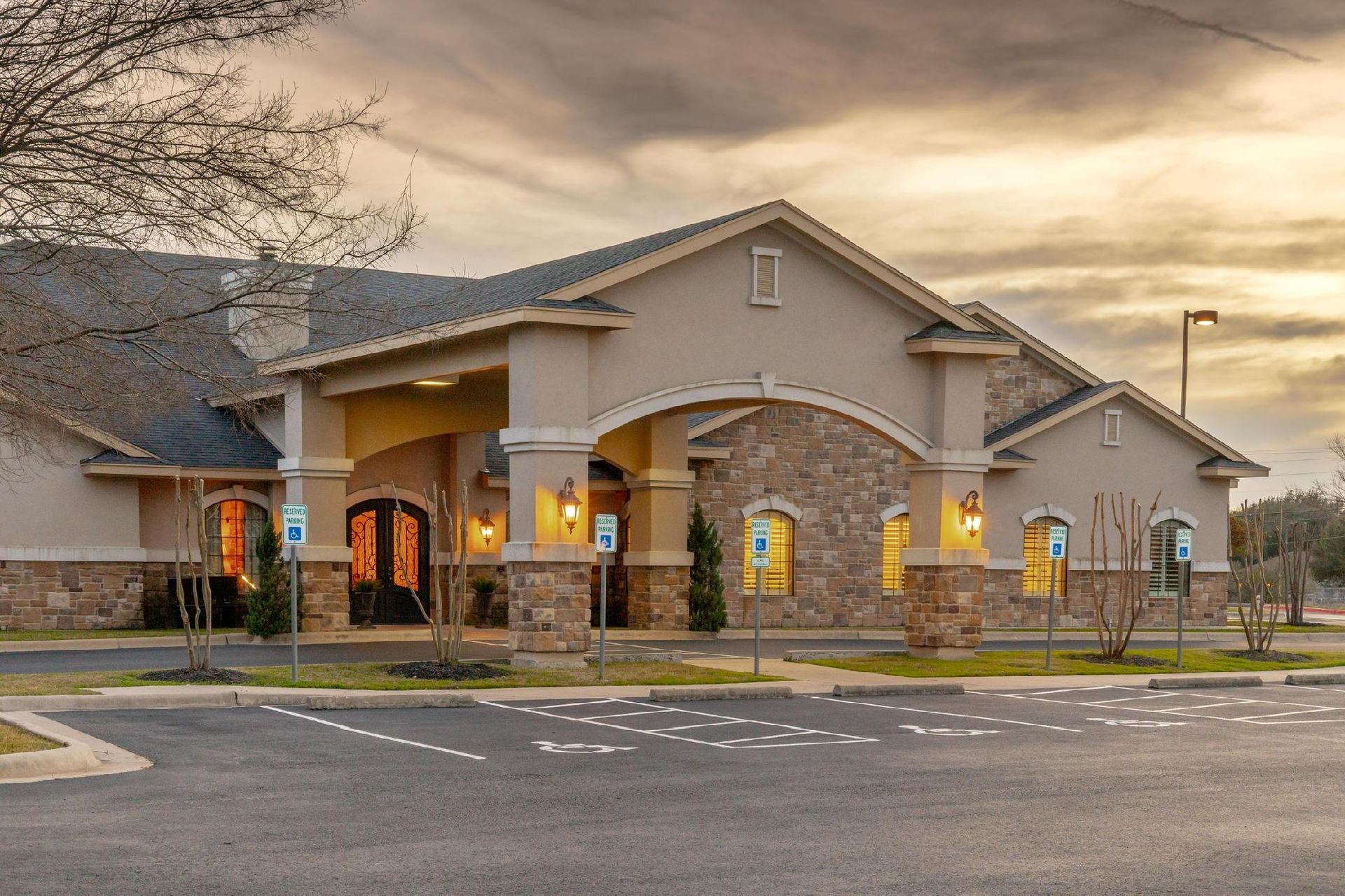 Beige building with stone accents, arched entrance, and parking spots. Cloudy sunset.