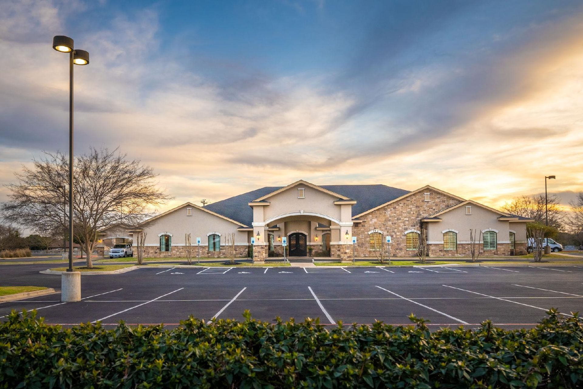 A single-story building with a parking lot at sunset; brown roof, beige walls, and stone accents.