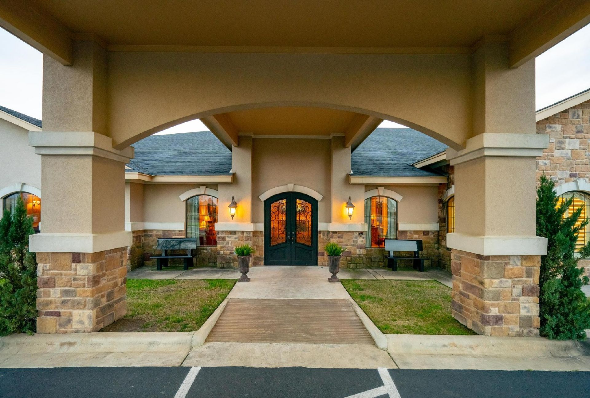 Entrance to a building with stone and beige accents, under a covered archway. Includes benches and double doors.