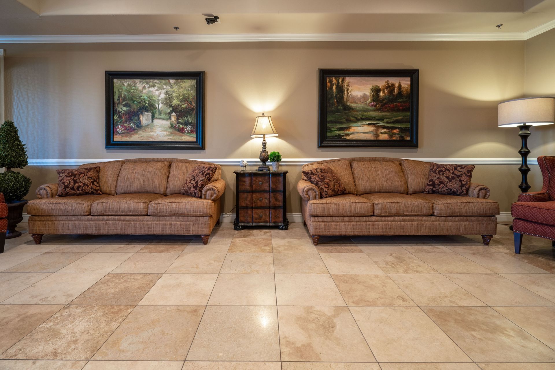Lobby with two brown sofas, a table with a lamp, paintings, and a tiled floor.