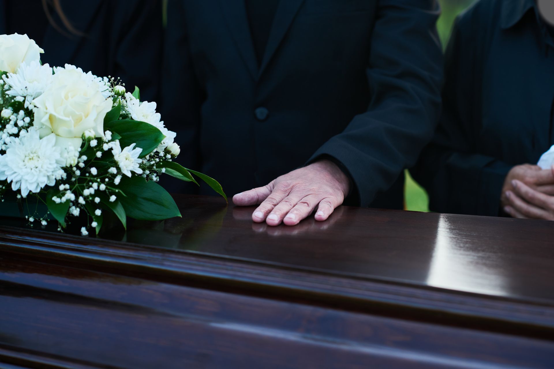 Person's hand resting on a closed, polished wooden casket next to a white flower arrangement at a funeral.
