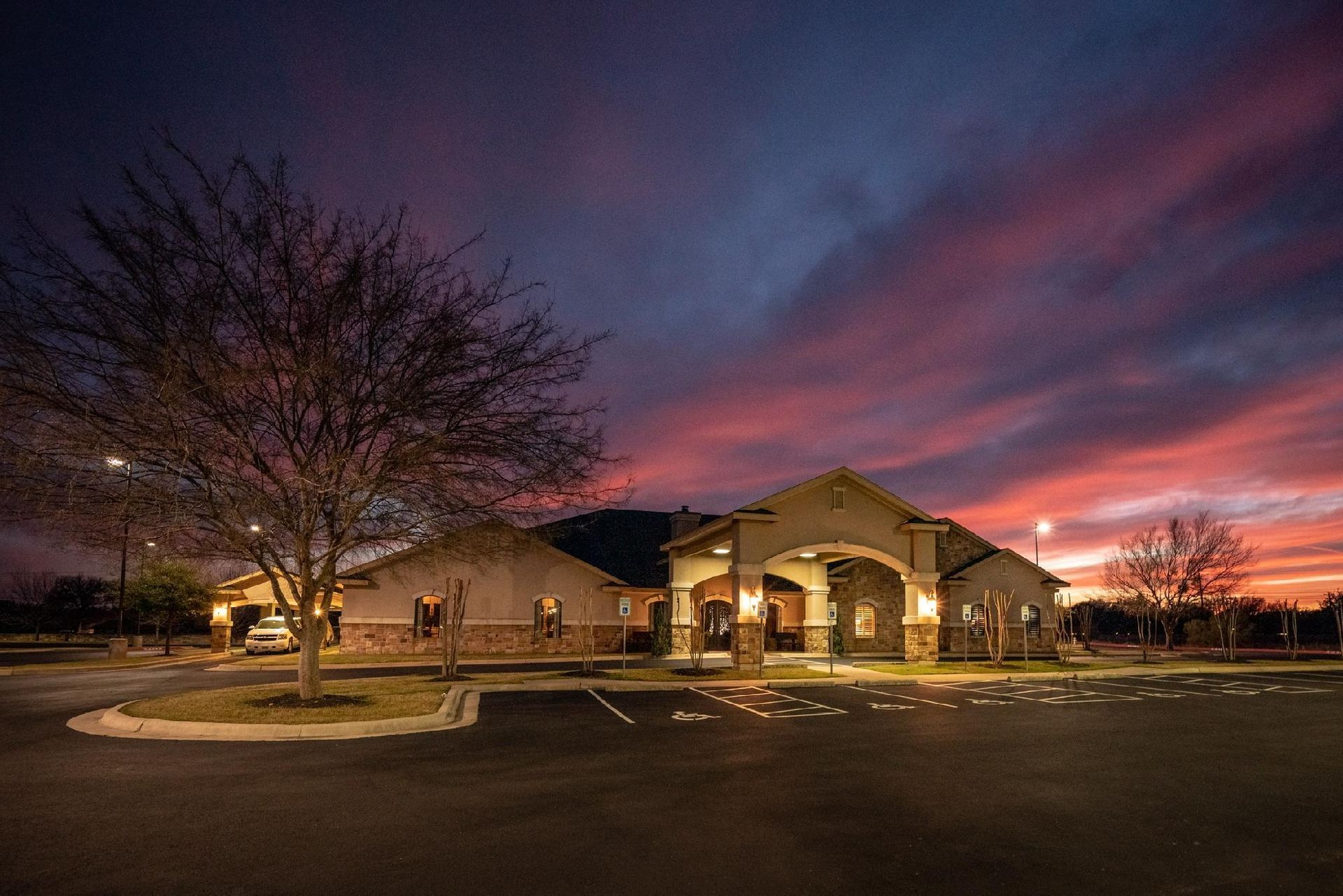 Building exterior at sunset with parking lot and tree. Sky is shades of pink, orange, and purple.